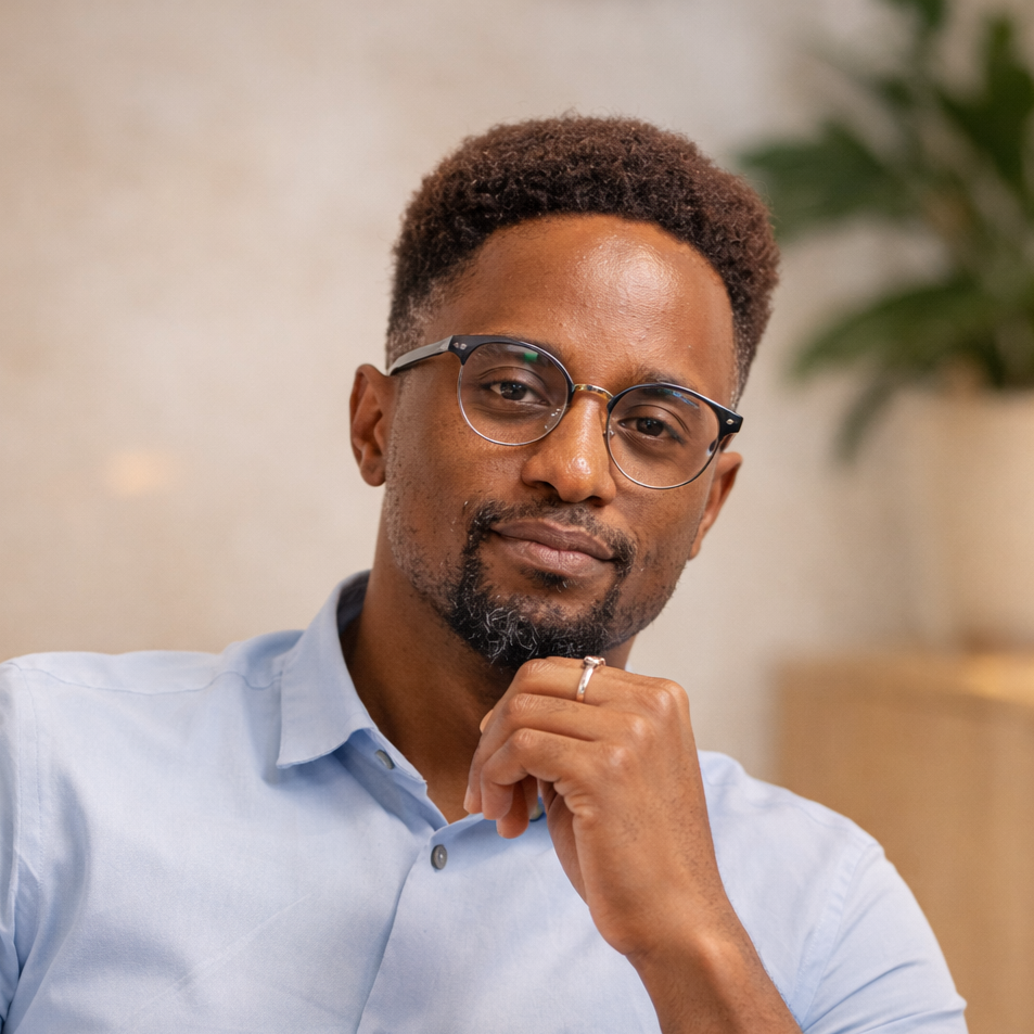 A man with glasses and a beard, wearing a light blue collared shirt, is sitting indoors with a neutral expression, hand under his chin, background with blurred indoor plants.