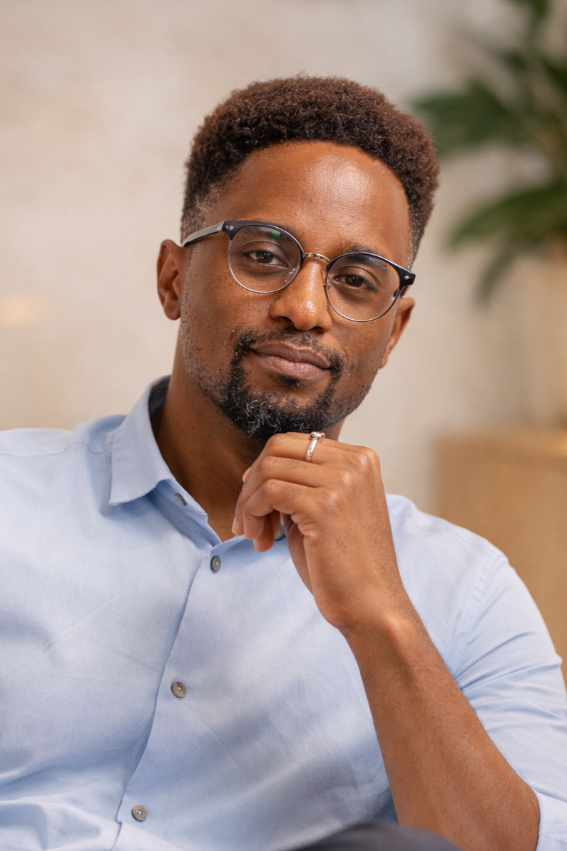 A man with brown, short curls, glasses, and a beard, wearing a light blue button-up shirt, resting his chin on his hand with a thoughtful expression, indoor setting with a blurred background and a green plant.