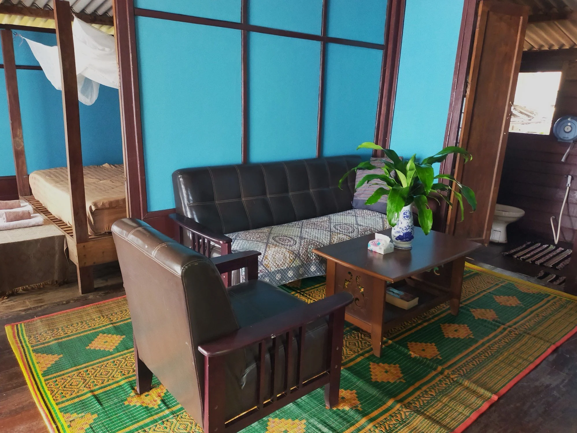 Living room with a black leather sofa, wooden coffee table with a potted plant, and a colorful woven rug on wooden floor, with a view of a sleeping area and a toilet in the background.