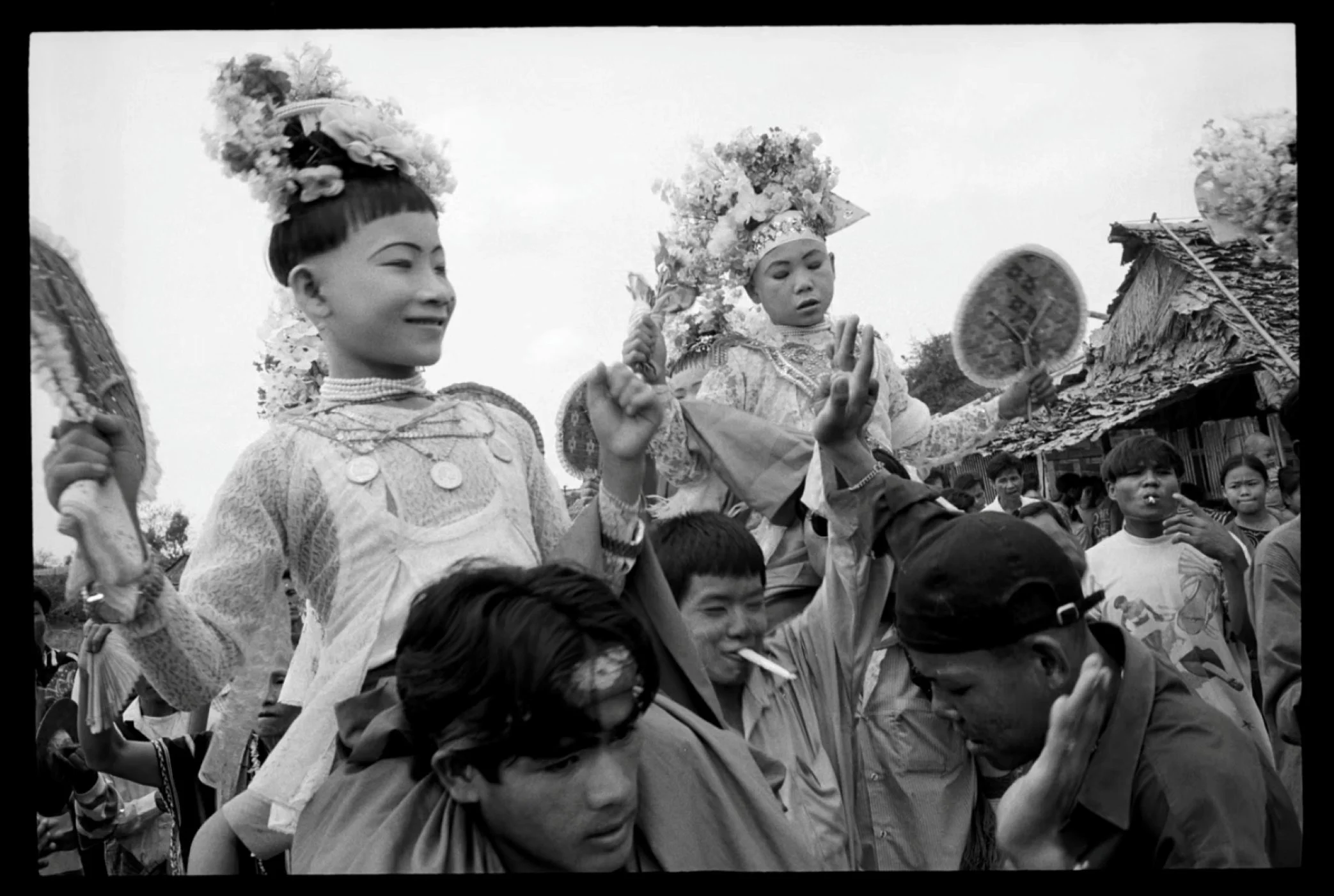 Young children dressed in traditional clothing, wearing headpieces adorned with flowers, are being carried on shoulders during a cultural festival or celebration. Several people, including children and adults, are visible in the background, some with expressive faces and colorful attire.
