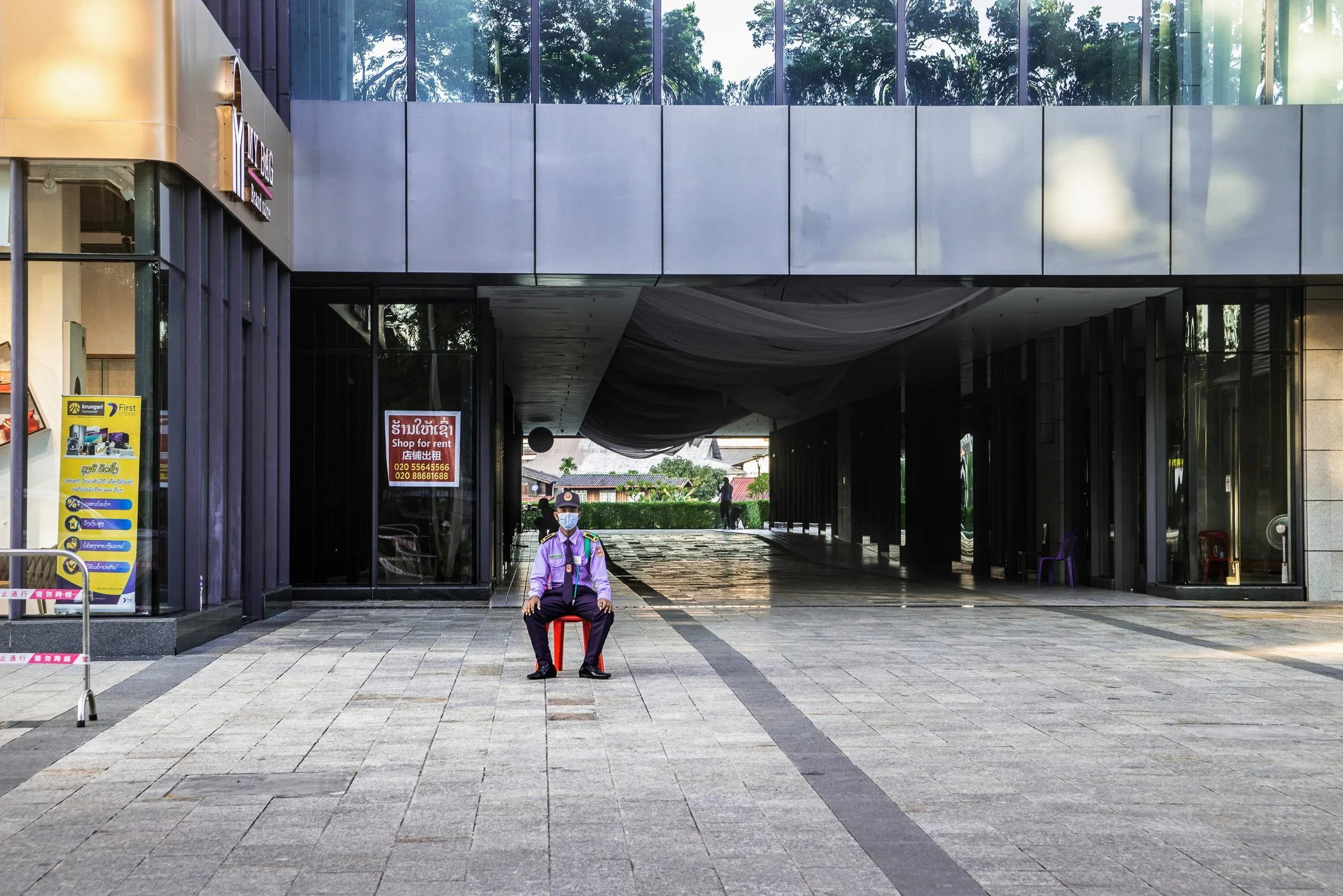 Vientiane, Laos, 2022 - A security guard sits outside a commercial building where shop spaces stand empty. After years of economic slowdown, many guards in Laos are employed less to manage crowds or protect busy businesses than to watch over closed s