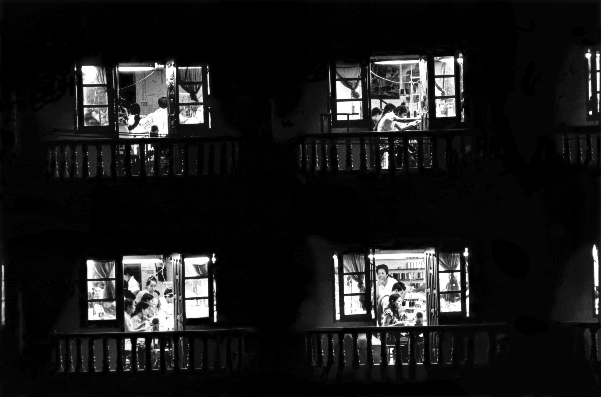 Black and white photo of a building with four open windows, each showing people inside. The interior appears to be a classroom or community space with children and adults engaged in activities.