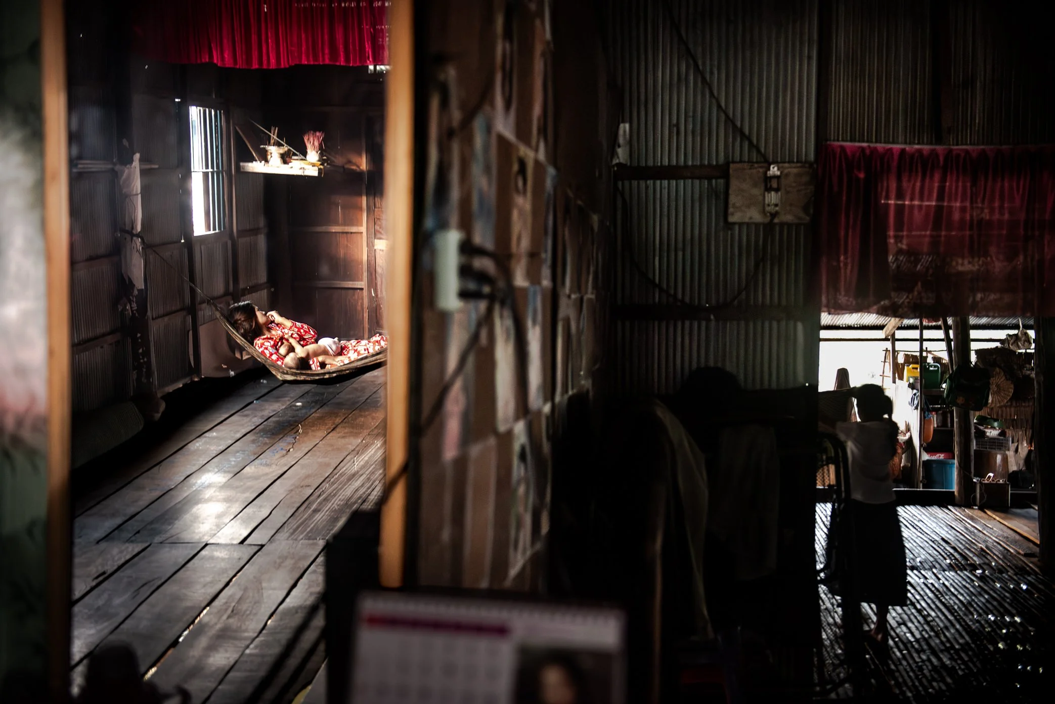 In the early morning inside a stilted home in Kampong Phluk, the wife of a fisherman waits for her husband to return from a night on the Tonle Sap. Their daughter prepares for school nearby, moving through the same quiet hours when the household wait