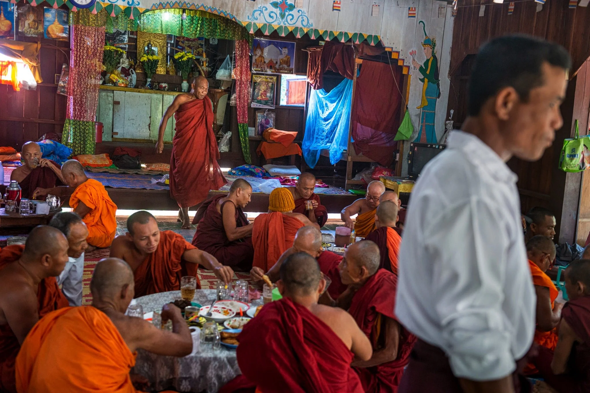 Group of Buddhist monks and a man gathered indoors, some seated at tables with food and drinks, others standing or walking, in a decorated room with religious artifacts and colorful banners.