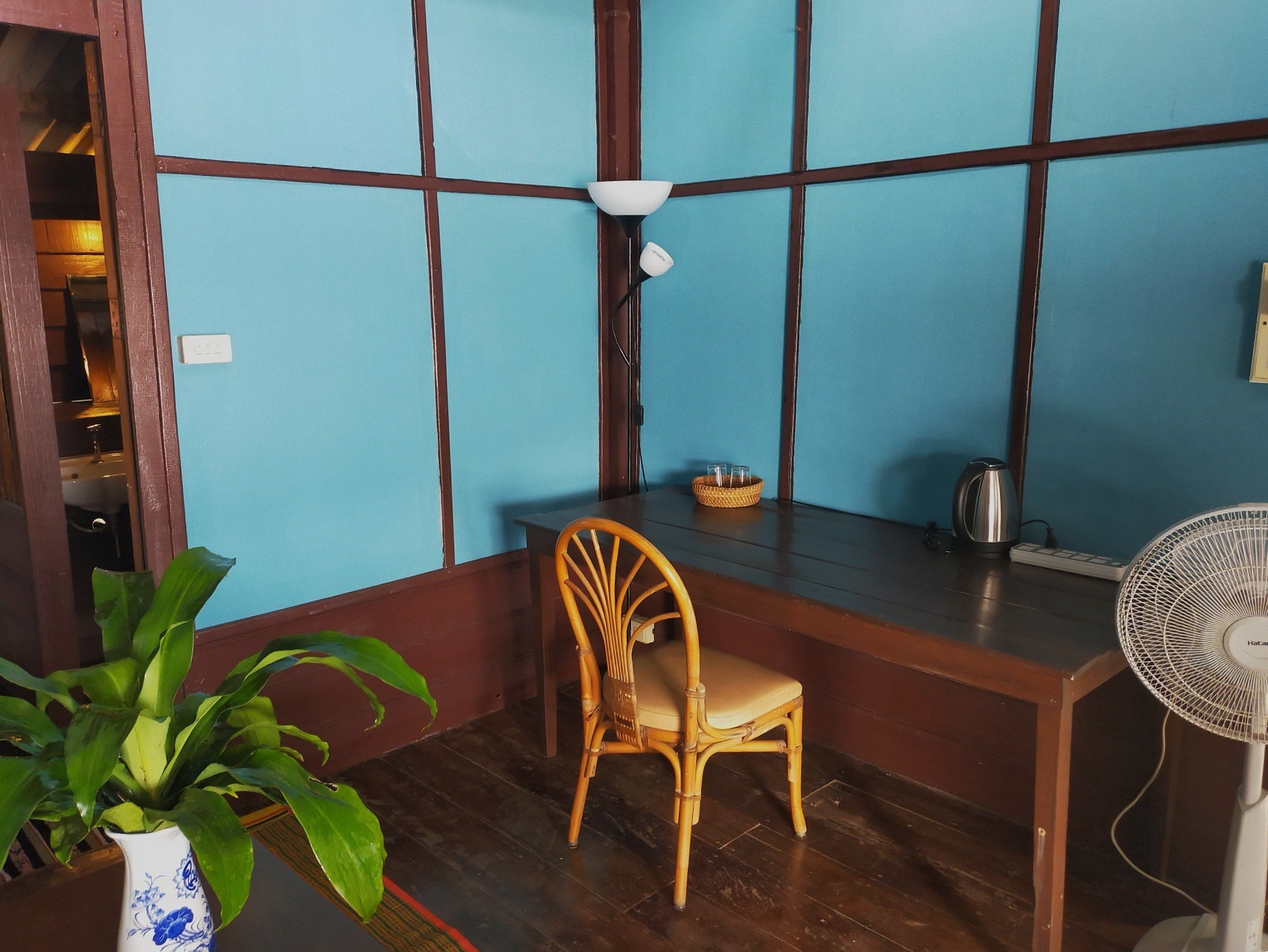 A small corner of a room with a wooden floor and blue-paneled walls. There is a woven armchair with a cushioned seat in front of a dark wooden table, which has a basket with cups and a silver electric kettle. To the right is a white oscillating fan, and in the foreground, a green leafy plant in a blue and white ceramic pot.