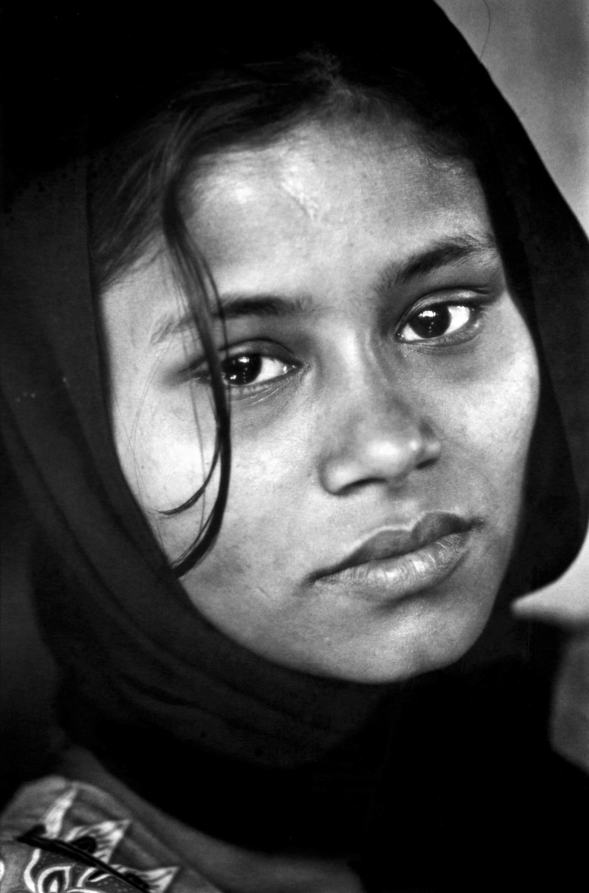 Close-up black and white portrait of a young woman with dark hair, wearing a headscarf, looking directly at the camera.