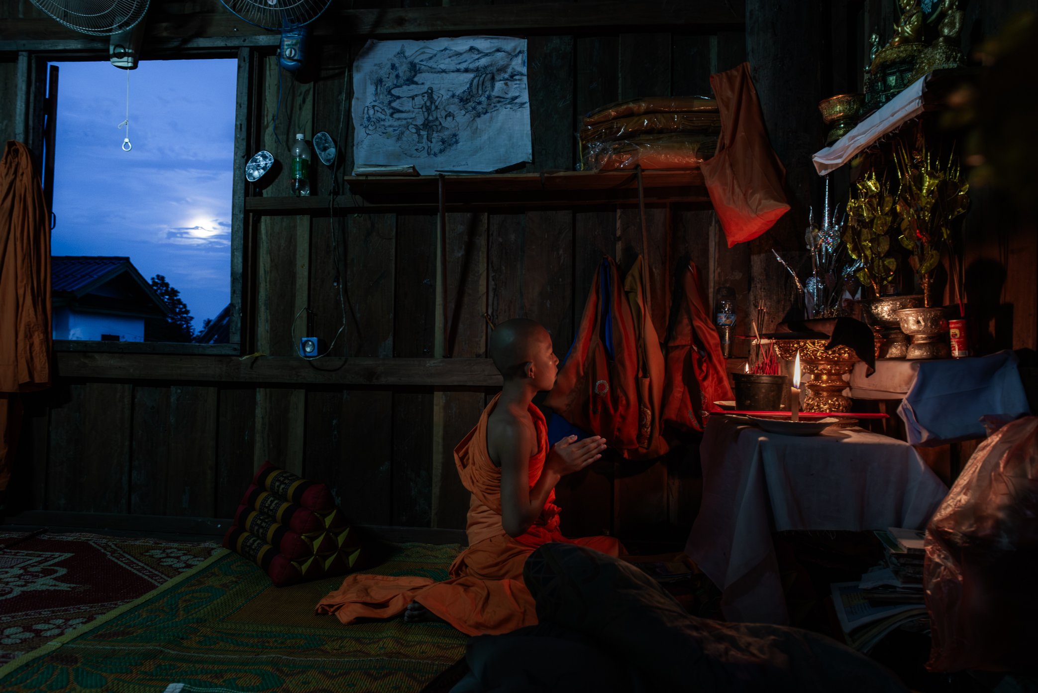 Inside a small wooden temple room in Don Khon, a young novice monk prays by candlelight as dusk settles over the Mekong. On the wall above him, a hand-drawn image of the local waterfalls reflects a landscape that shapes both faith and livelihood here