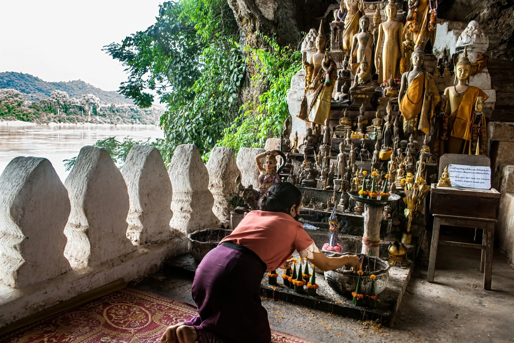 Pak Ou Caves, Luang Prabang. Laos, 2022 - At Tham Ting, one of the sacred Pak Ou caves overlooking the Mekong River, a souvenir seller makes an offering and prays for better business. The caves, among Luang Prabang’s best-known pilgrimage and tourist