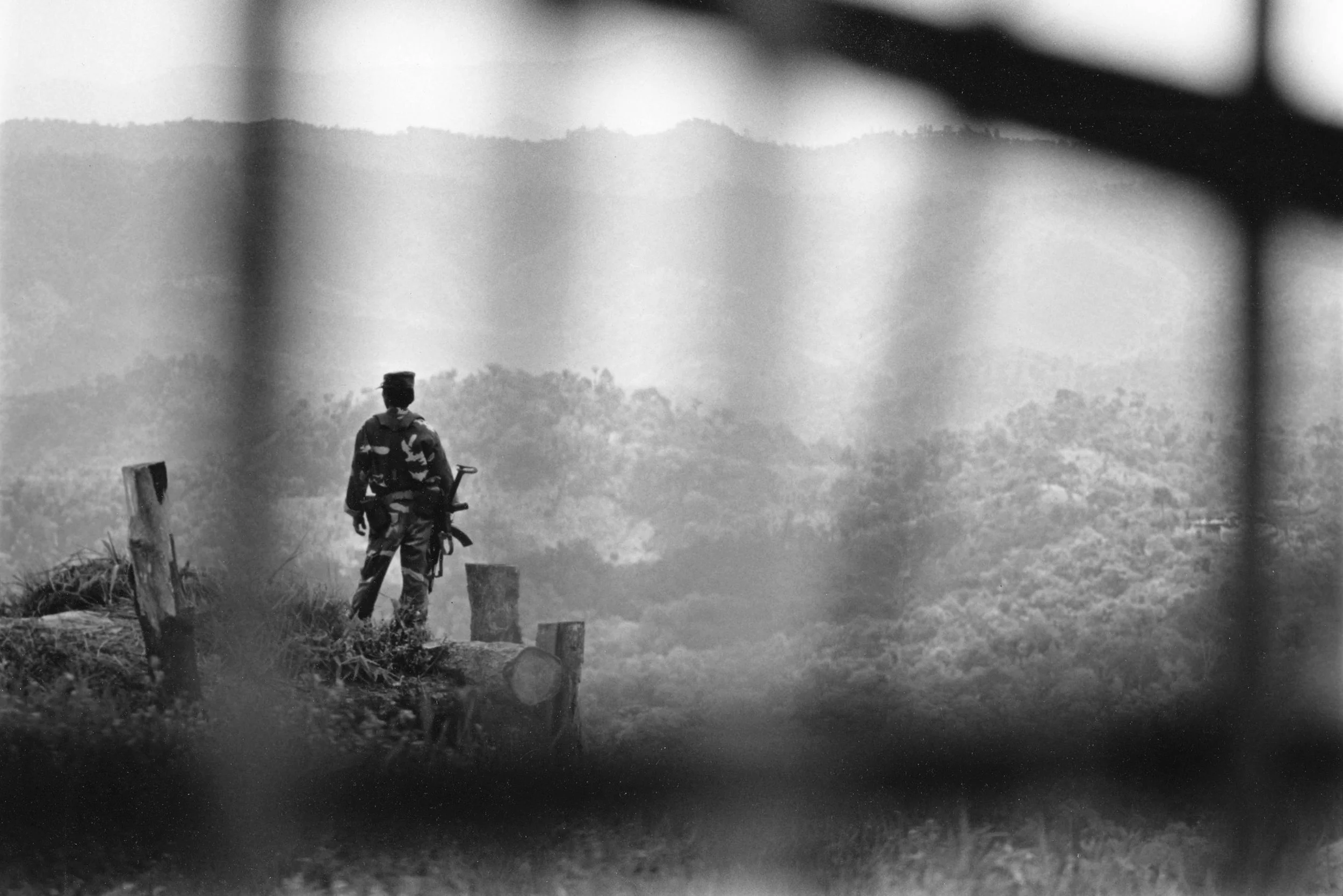 A soldier standing on a fence overlooking a hilly landscape, holding a rifle, viewed through a window or bars.