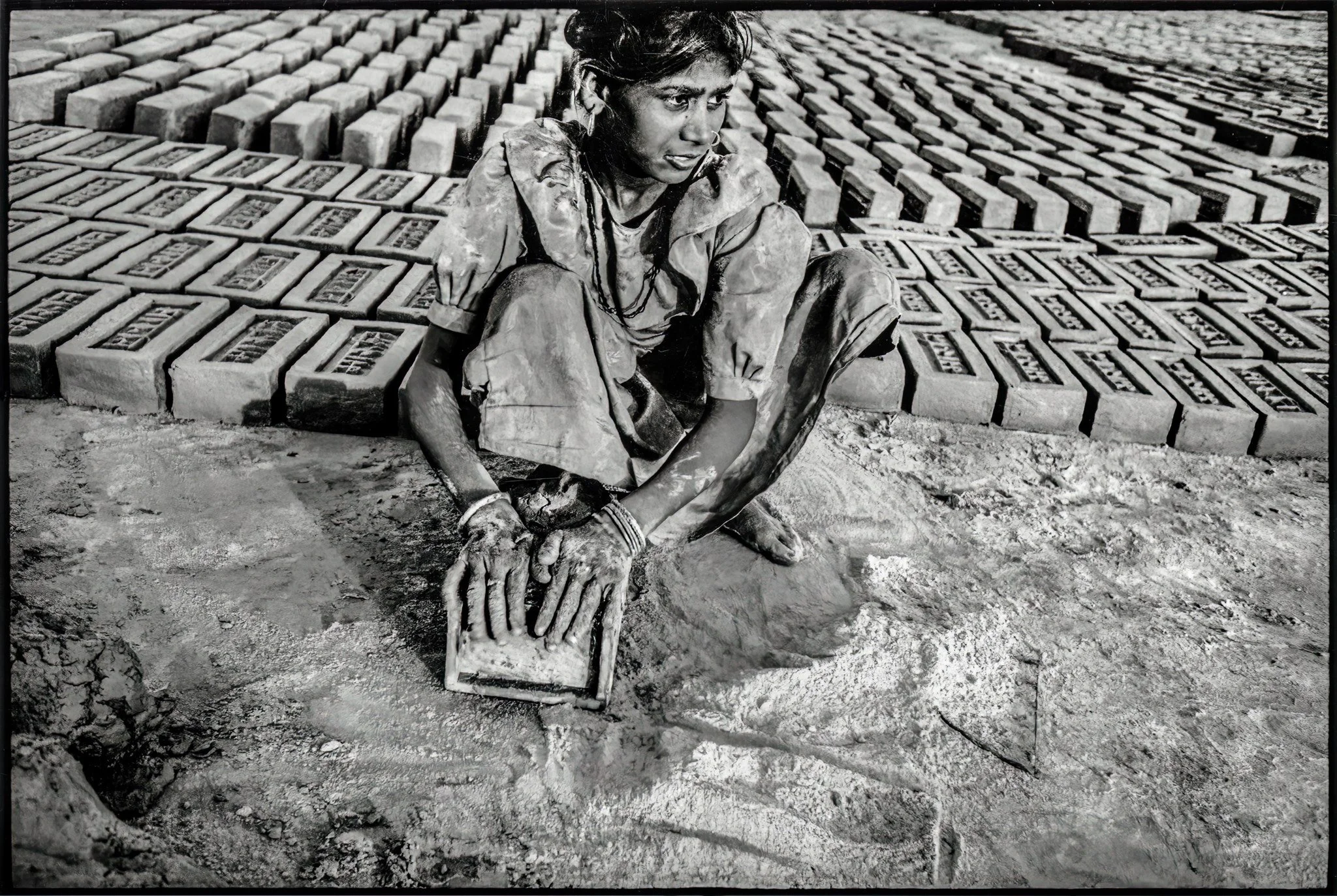 At a brick kiln - Patna, in the Indian state of Bihar, a child shapes wet clay by hand before it is left to dry in the sun. Rows of newly made bricks stretch across the yard behind her, each one carrying the mark of human labour before it enters the 