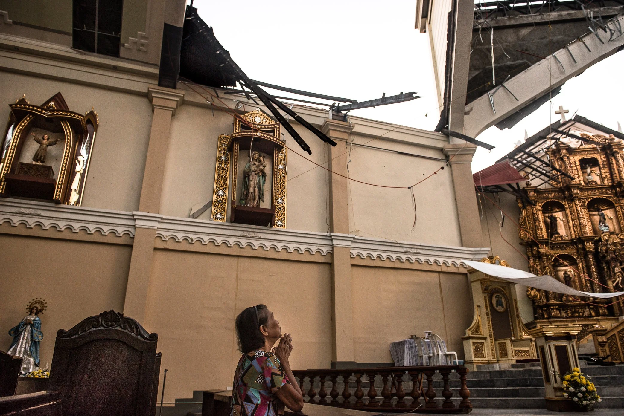 Palo, Leyte, Philippines, 2013 - A woman prays inside a church damaged by Typhoon Haiyan. Around her, the roof has been torn open and sacred figures remain exposed beneath the broken structure - a quiet image of faith held in place amid loss, ruin, a
