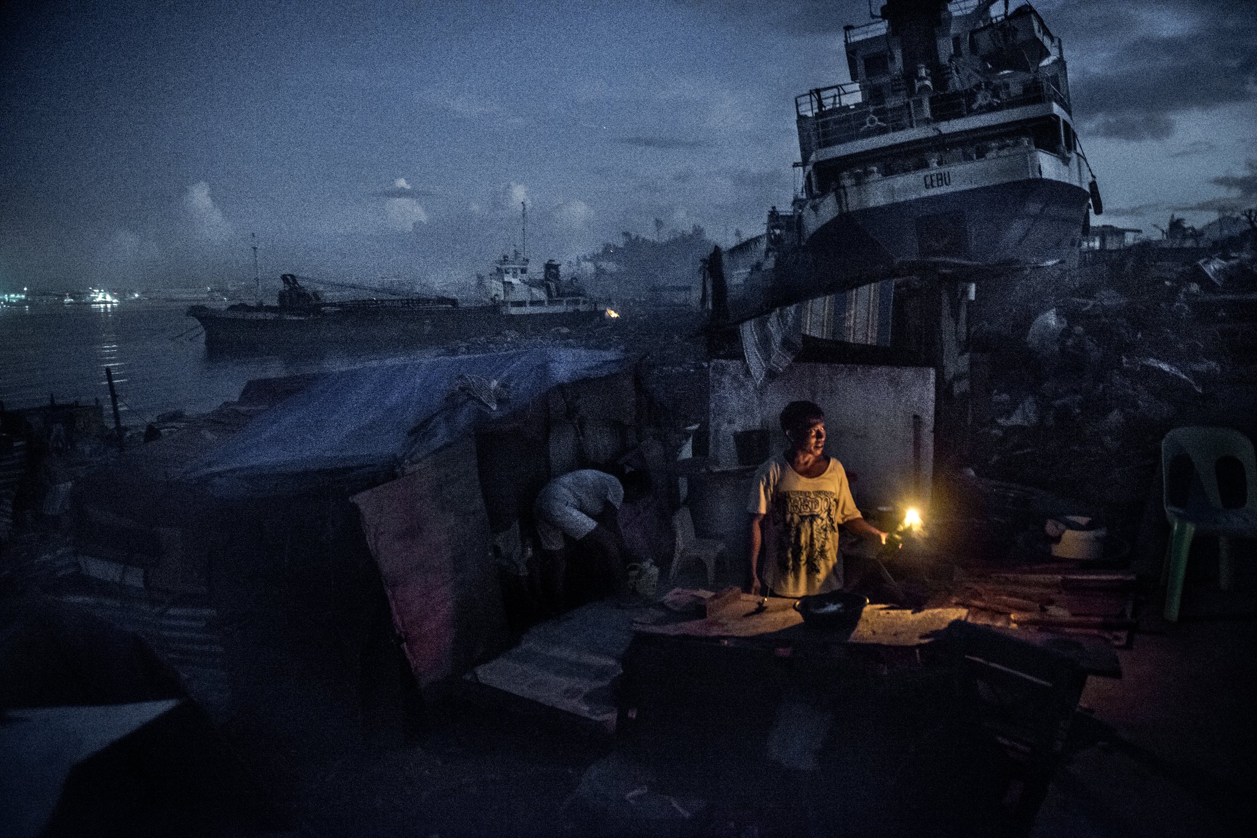 Person holding a yellow light standing near damaged boats and debris at a dock during nighttime.
