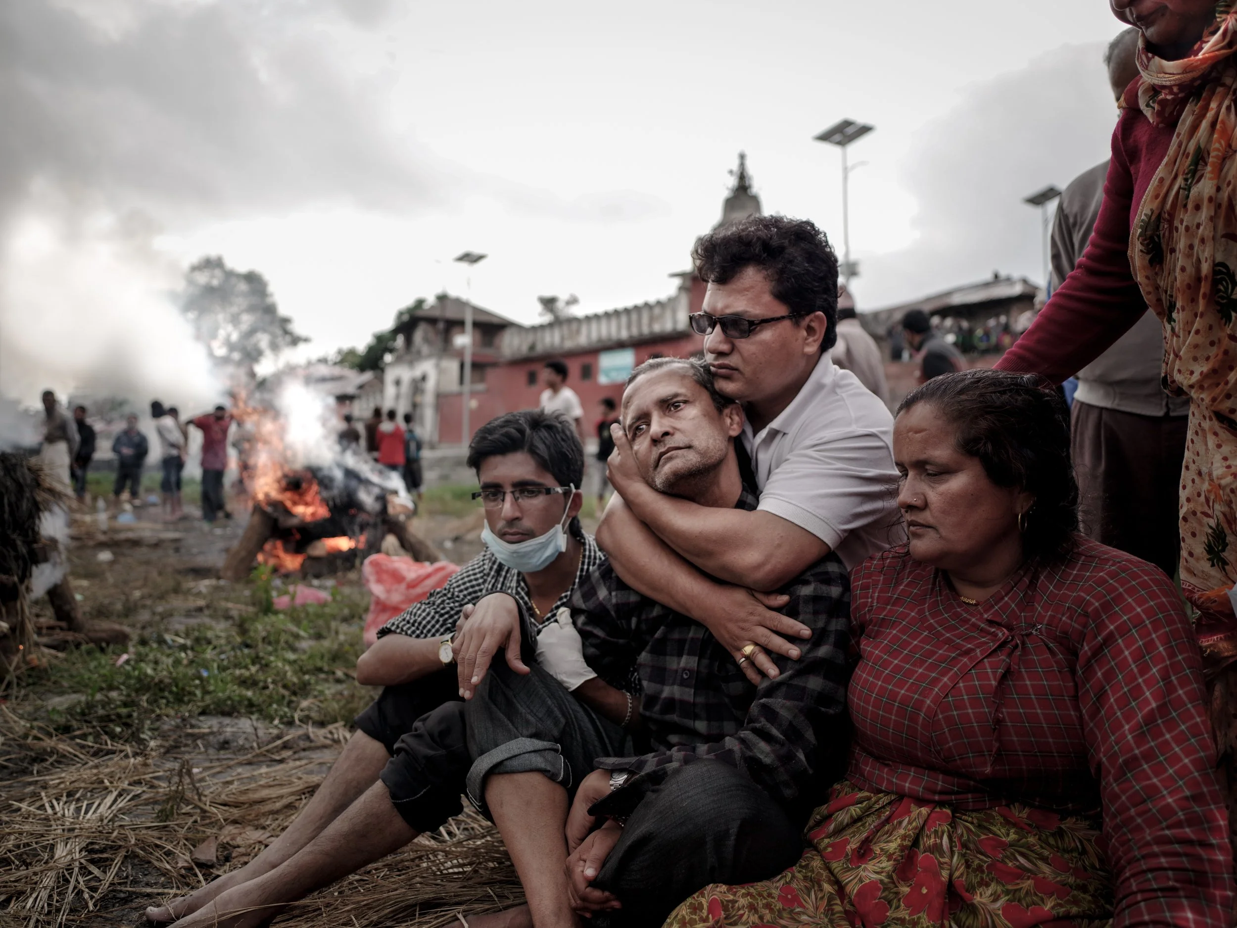 Group of people sitting and mourning near a burning pyre outdoors during cloudy weather.