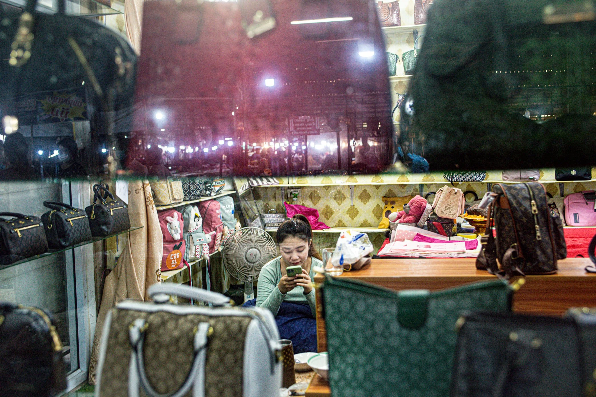 View through a shop window at night showing a woman sitting at a cluttered table, looking at her phone, surrounded by handbags and bags, with city lights and street scene reflections visible in the glass.