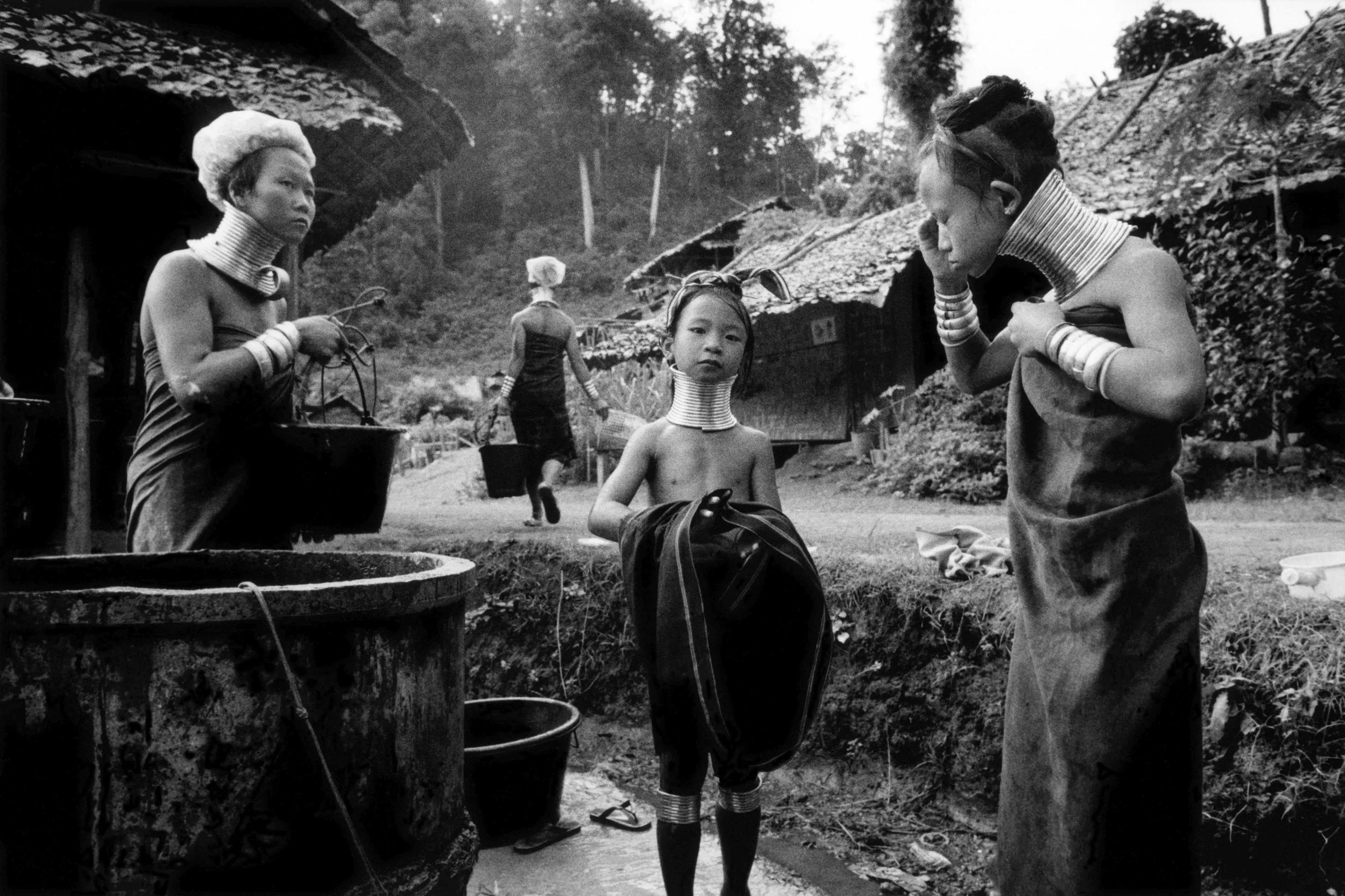 Traditional women from a Southeast Asian hill tribe, adorned with metal neck rings, standing around a water well, with rustic houses and trees in the background.