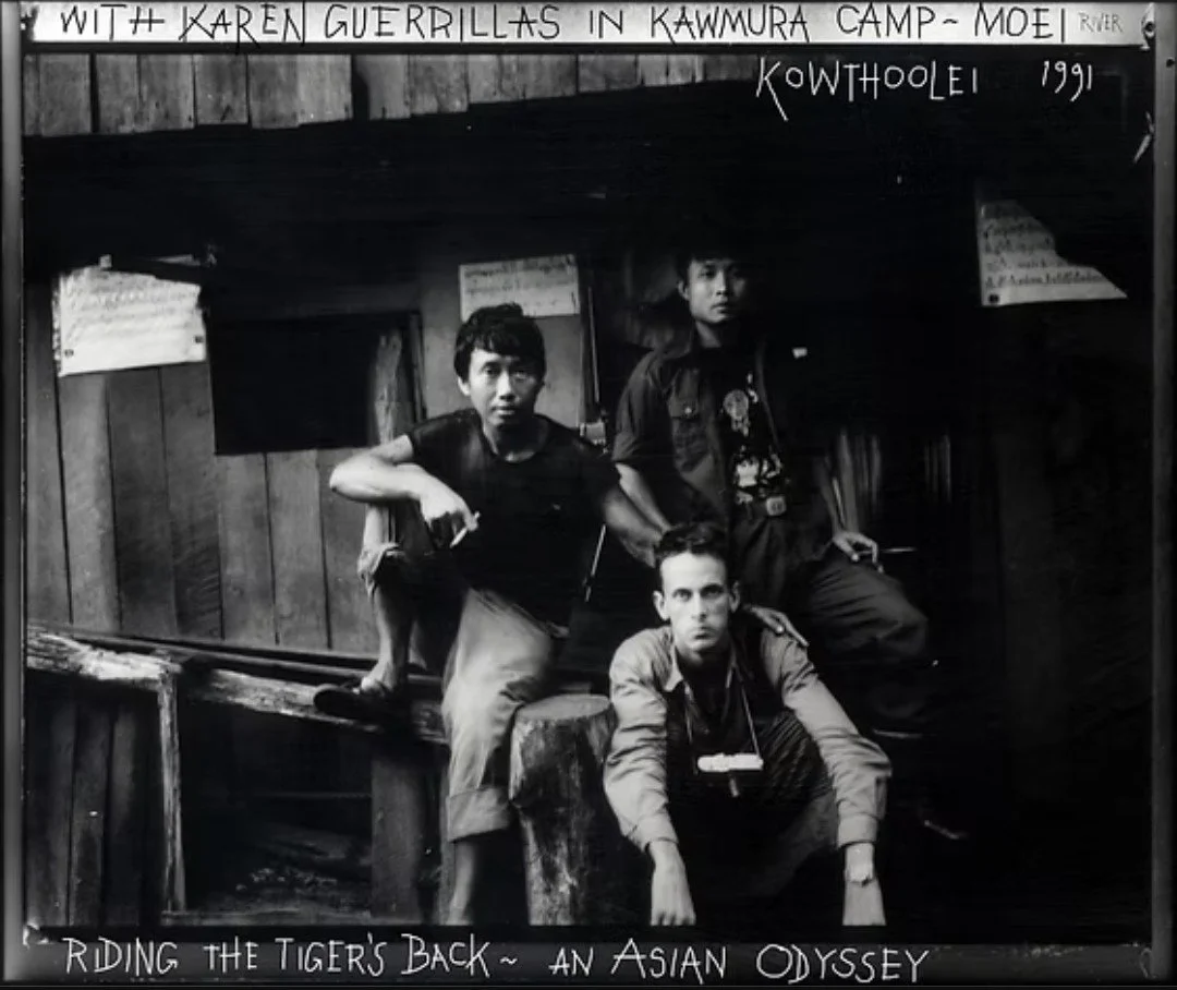A black and white photo of three young men inside a wooden structure. The man on the left is sitting on a wooden beam with one arm resting on his knee. The man on the right is standing with one leg up on a wooden platform. The man in the front is crouching down with both hands on the floor. There are signs and writings on the wall behind them, including "With Karen Guerrillas in Kawmura Camp - Moei" and "Riding the Tiger's Back - An Asian Odyssey." The photo appears to be from 1991 and has a caption noting a connection to Kowthoolei.