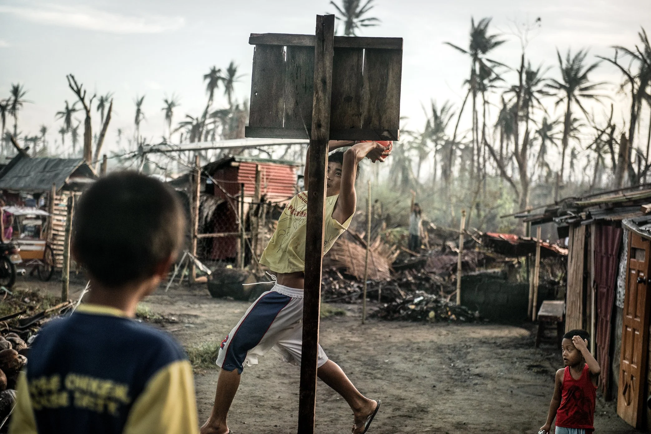 Tacloban, Philippines, 2013 - Boys play basketball with a makeshift hoop in a neighborhood devastated by Typhoon Haiyan. Around them, homes are damaged, trees stripped bare, and the landscape still carries the force of the storm. Yet in the middle of
