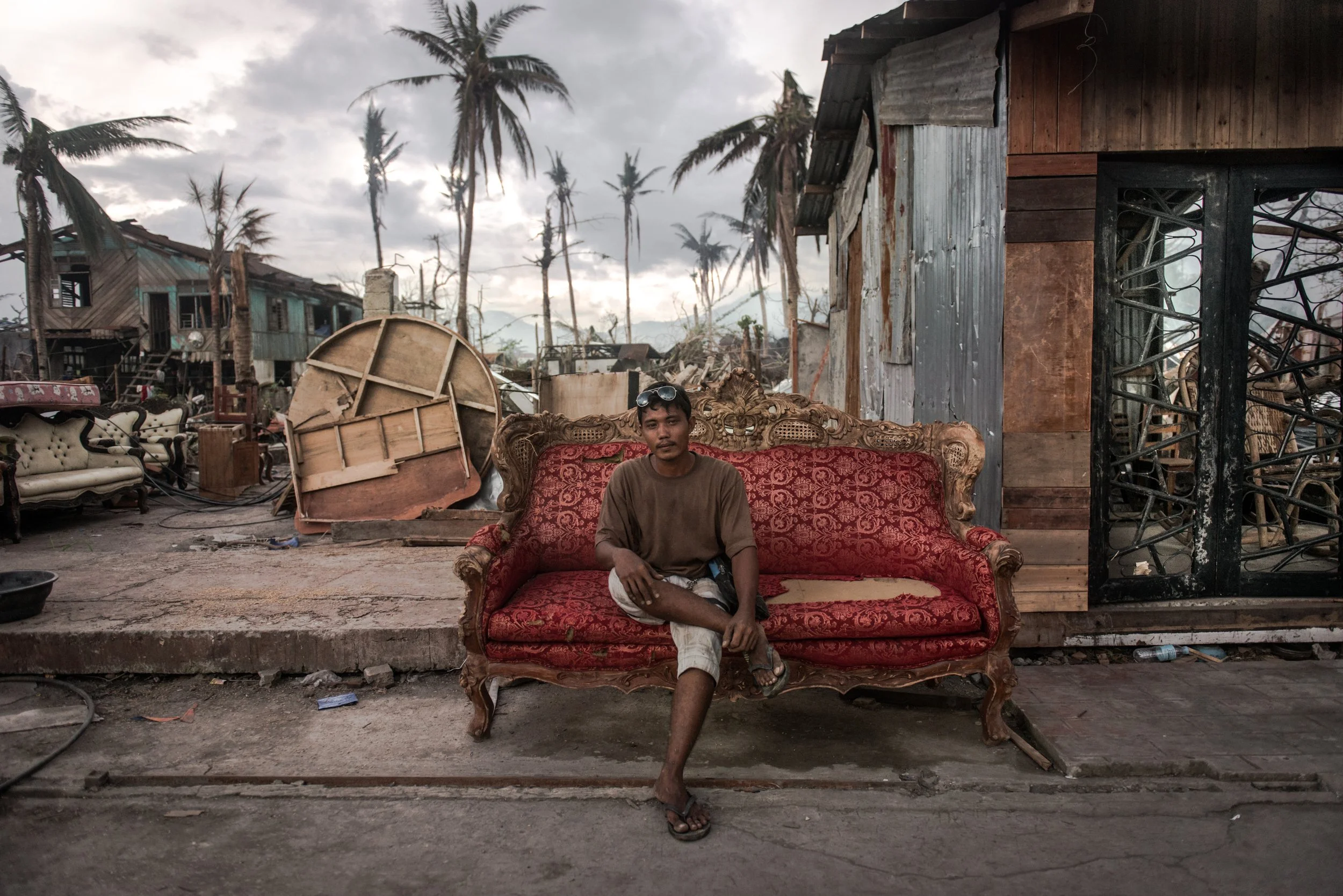 A young man sits on a vintage red sofa in a damaged neighborhood with debris and damaged houses around him, under cloudy sky.