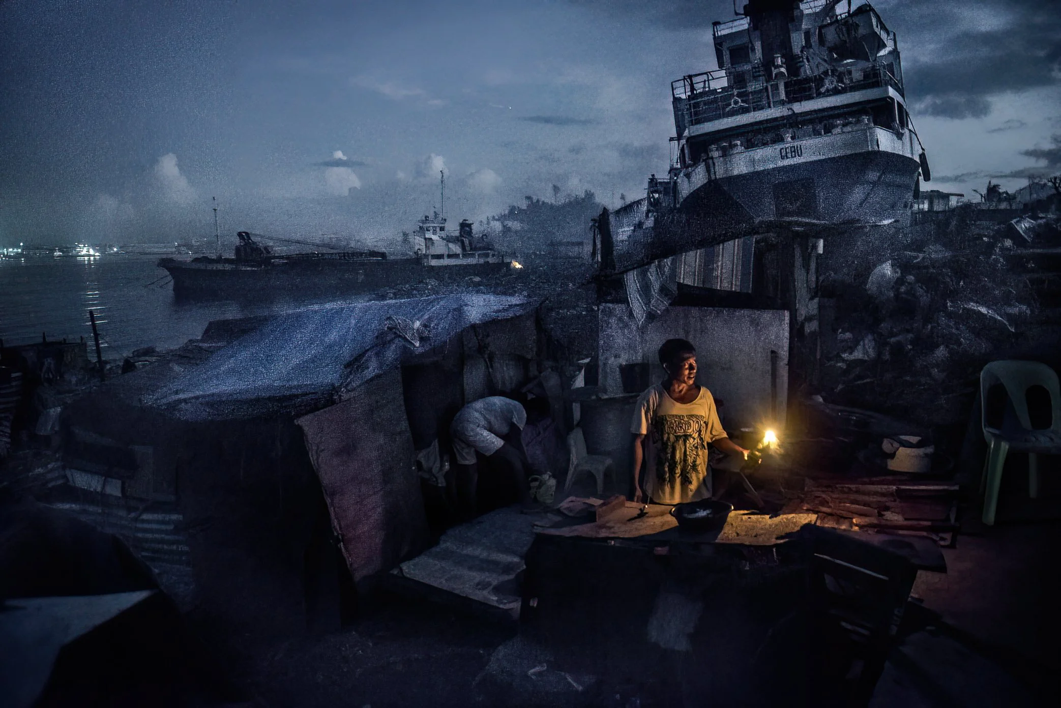 Tacloban, Philippines, 2013 - In the port area of Tacloban, ships driven ashore by Typhoon Haiyan loom over homes reduced to makeshift shelters. By candlelight, a man prepares food for his family, cooking beneath a surreal landscape of wrecked vessel