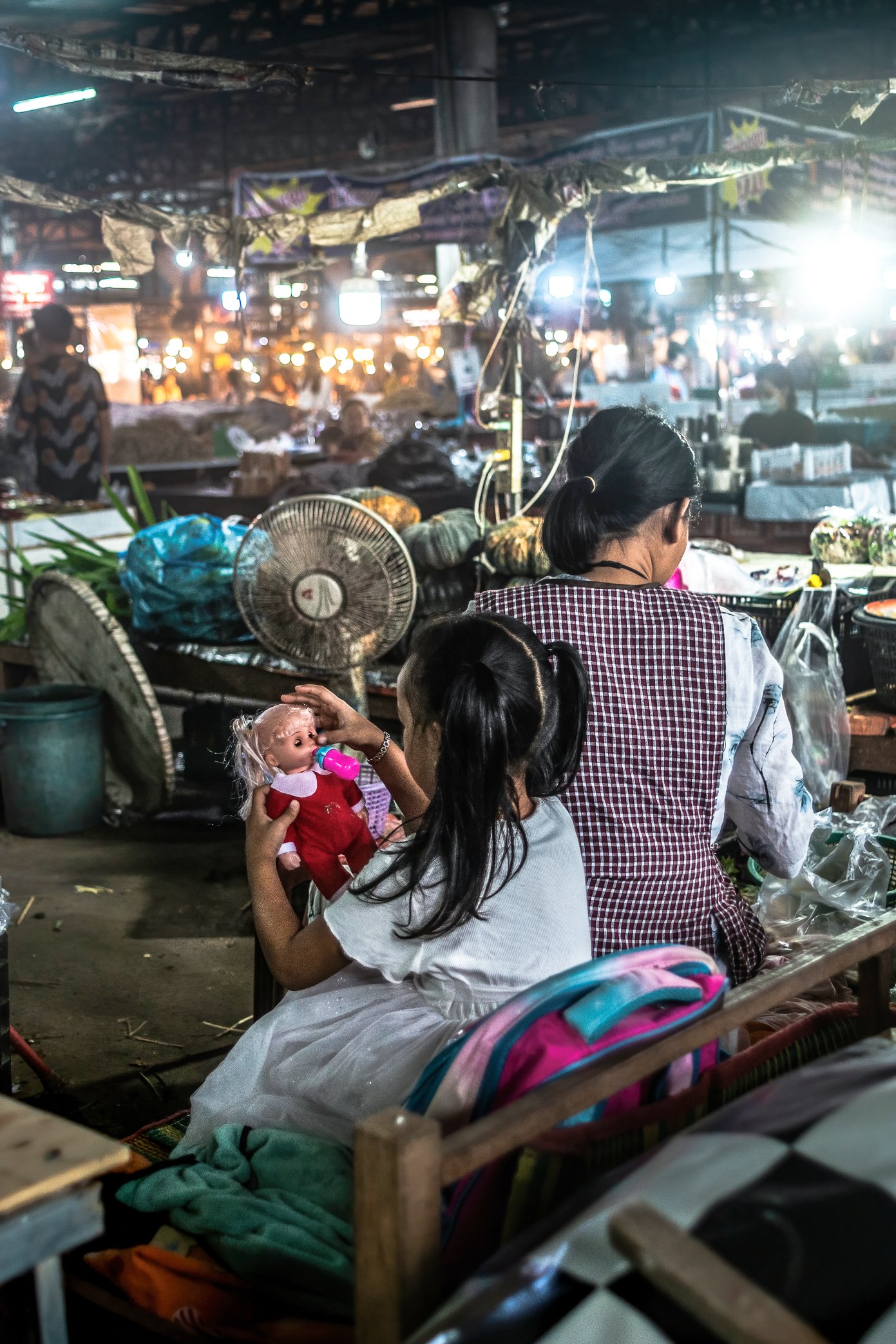 Vientiane, Laos, 2022 - A child plays beside her mother’s stall at Oxy wholesale fresh market. As purchasing power weakens and sales slow, many vendors spend longer hours waiting for customers, often bringing their children and family members with th