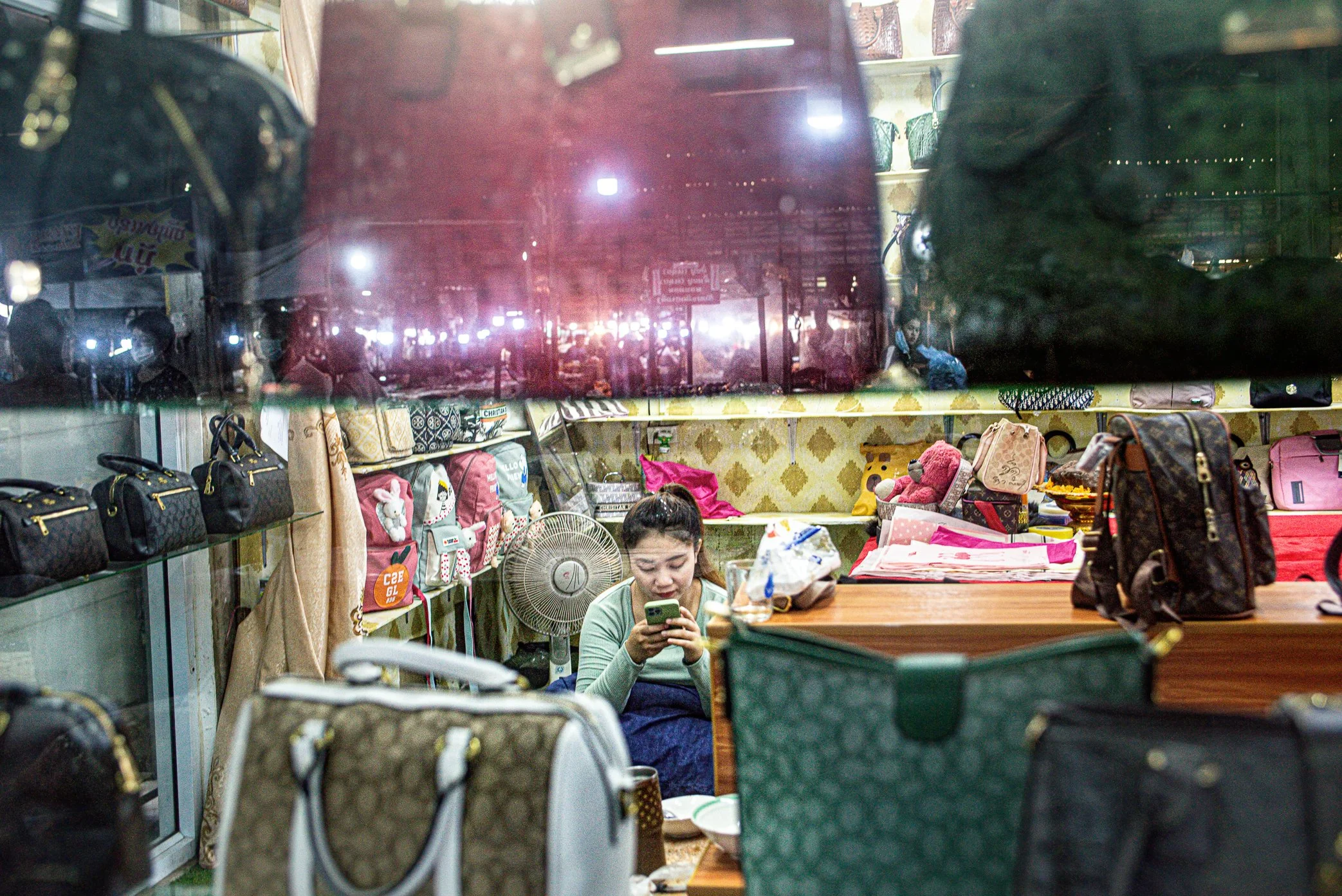 Vientiane, Laos, 2022 - A shopkeeper sits behind a display of copy-brand handbags and fashion goods as traffic lights reflect across the shop window. For many young Lao consumers, foreign fashion remains desirable, but purchasing power has weakened s