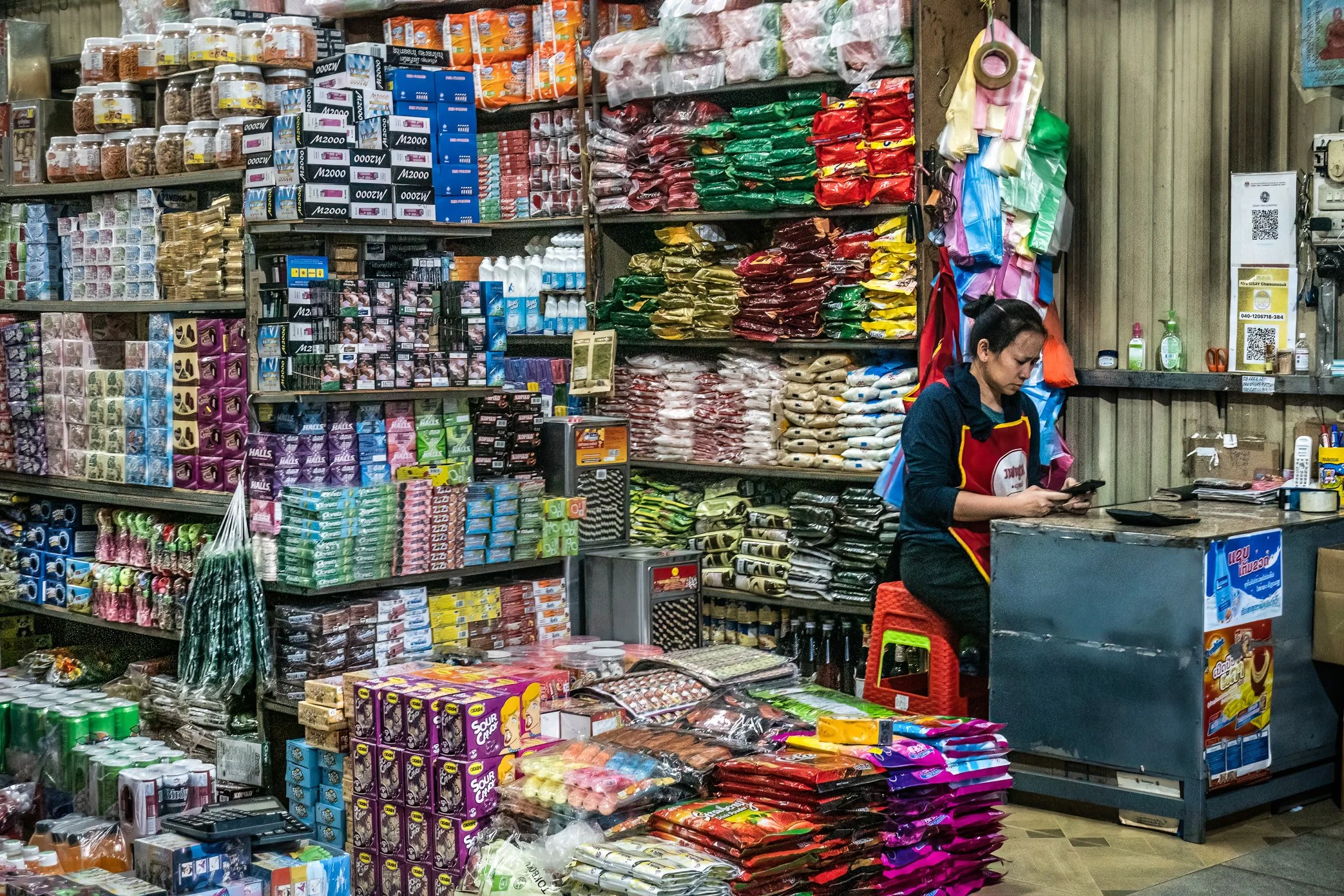 Pakse, Laos, 2022 - A shopkeeper waits among shelves of imported goods at a grocery stall in Pakse market. As the Lao kip weakens, prices for products brought in from neighboring countries become harder to predict, leaving small retailers uncertain a