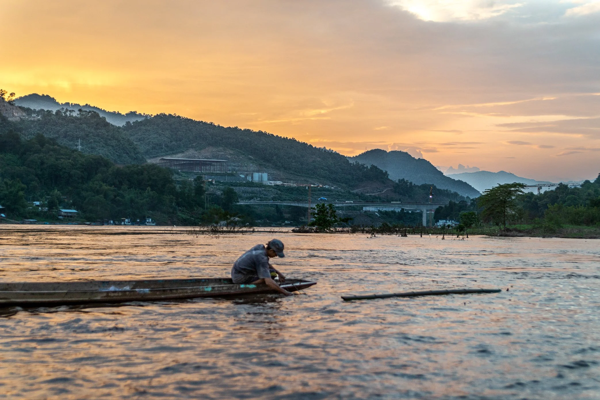 A fisherman from Pak Nam Ou works the evening water in front of his village as construction rises across the Mekong behind him. The bridge, built at speed through the mountain landscape, has become a sign of a larger transformation along the river.

