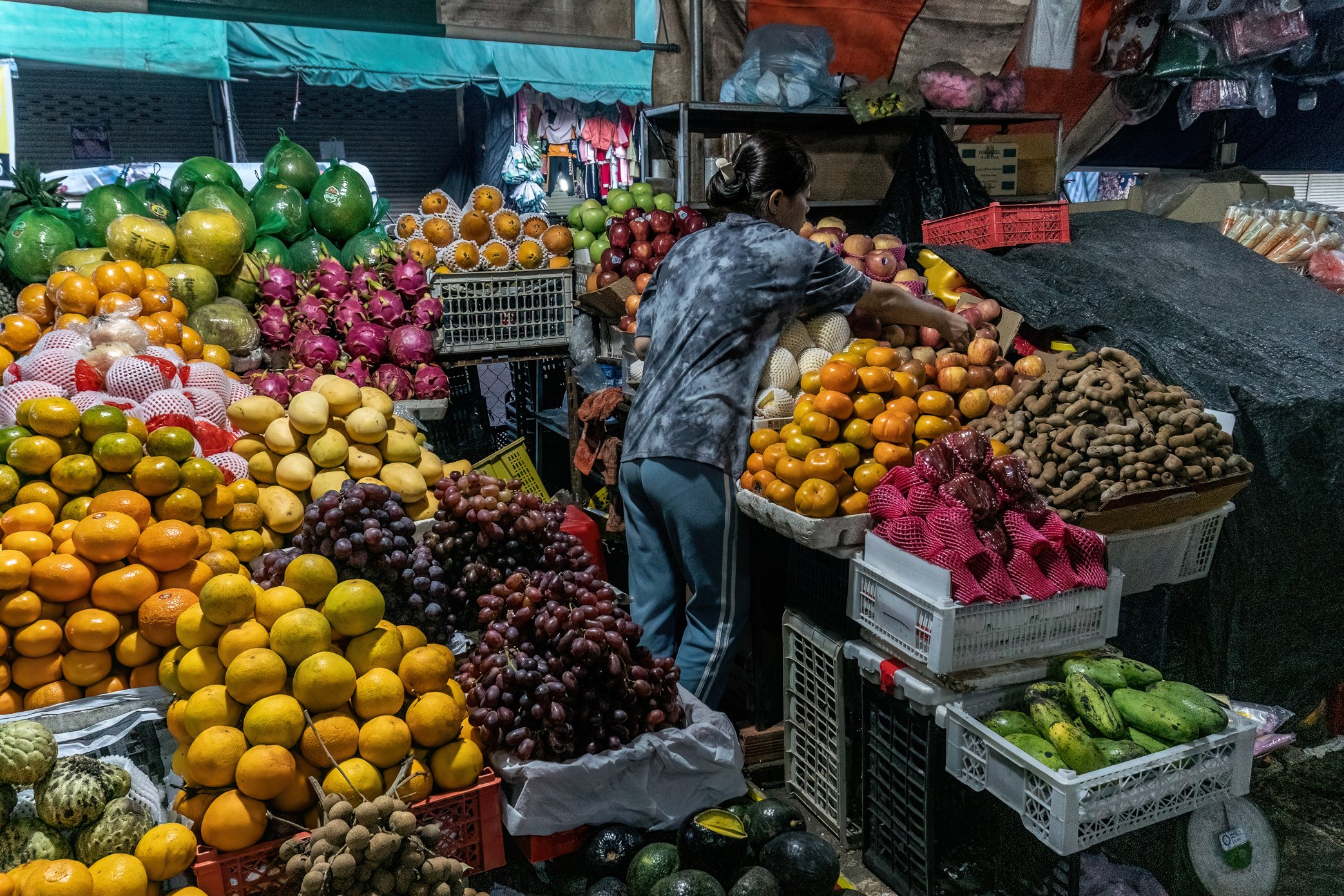Pakse, Laos, 2022 - A fruit seller prepares her stall before the market opens. Much of the fruit she sells is imported from countries with stronger currencies than the Lao kip, making each purchase more expensive before it even reaches her shop. As t