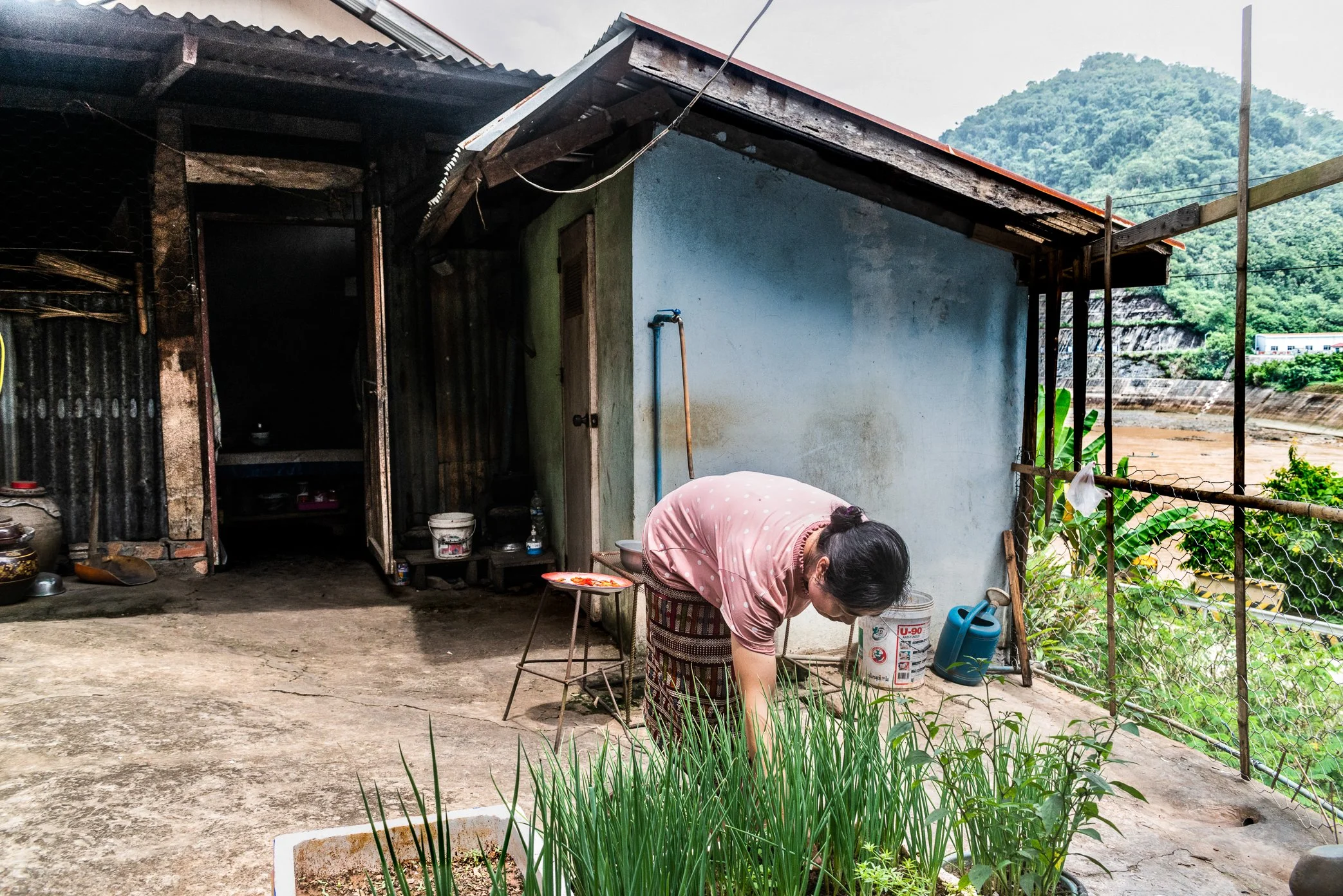 A woman tends a small vegetable bed beside her home along the Nam Ou River, where daily life has been reshaped by the construction and operation of hydropower dams.

Before the dam altered this stretch of river, the space behind many houses was part 
