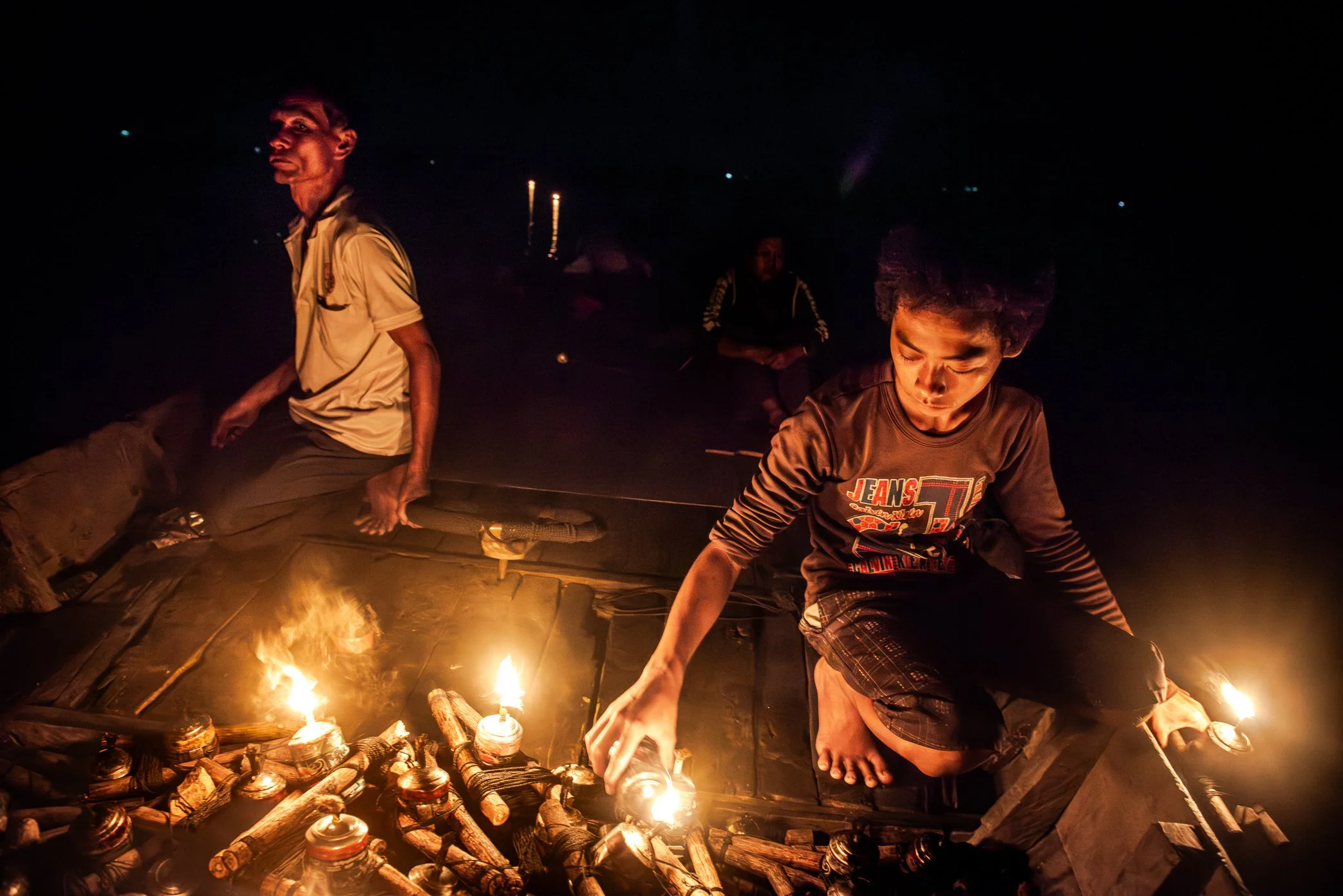 Tonle Sap, Cambodia - A young fisherman lights kerosene lamps before a night of fishing on the Tonle Sap. The flames will be set across the water to attract insects, drawing fish toward the glow. On boats like this, children often learn the work earl