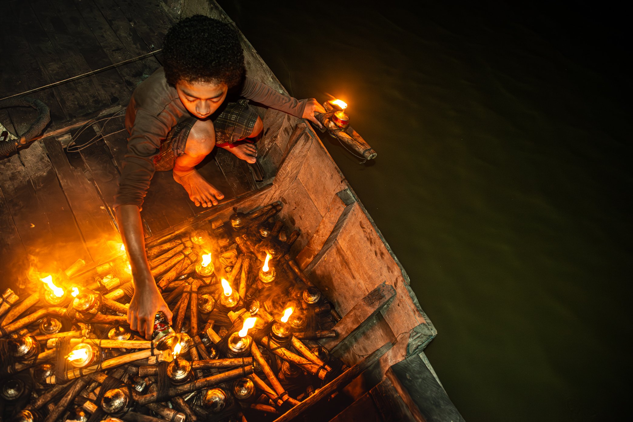 Tonle Sap, Cambodia - The boat owner’s son prepares to set a kerosene lamp onto the dark water of the Tonle Sap. Each flame is part of a night-fishing method used to draw insects and fish toward the boat. For families who live from the lake, children