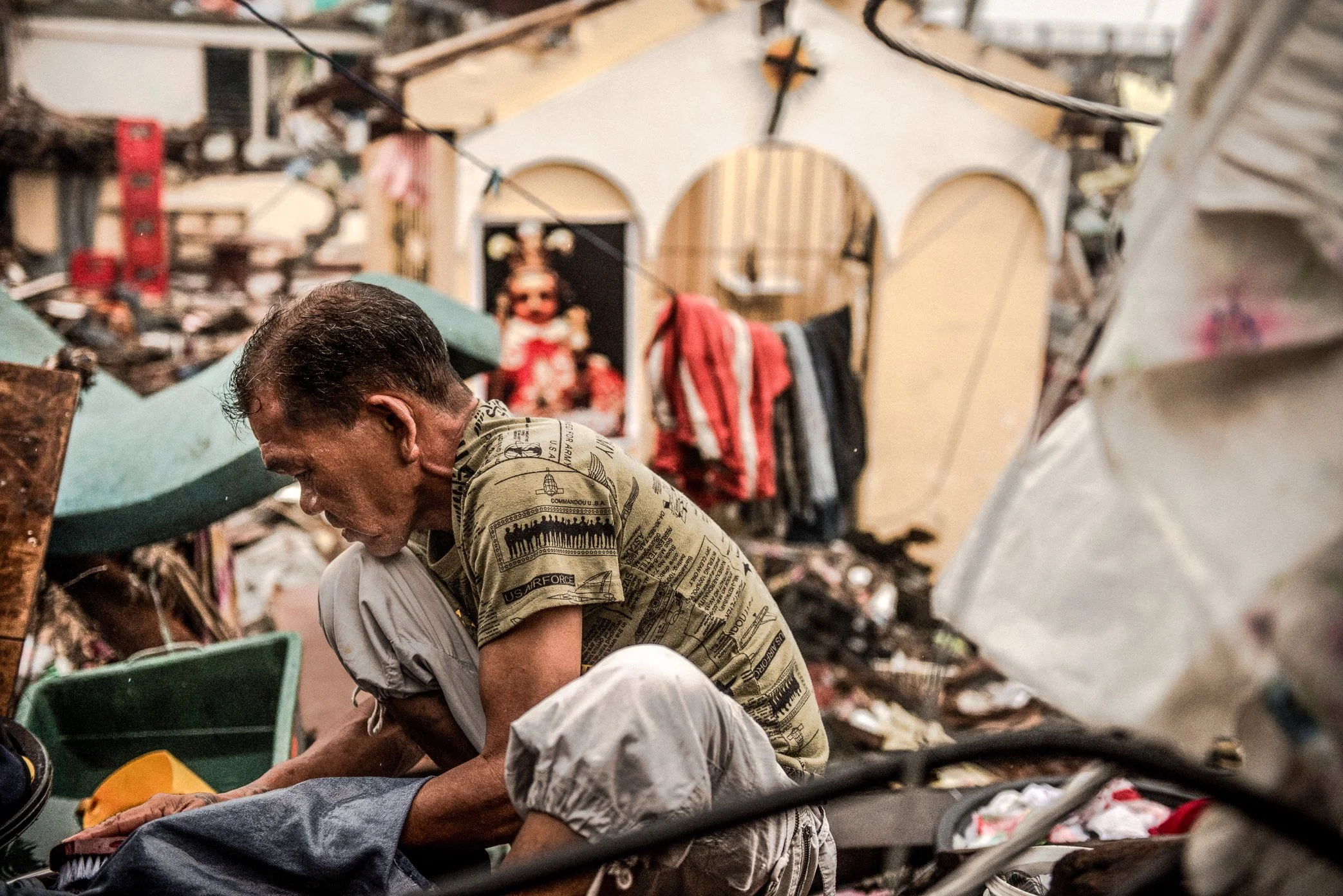 Leyte, Philippines, 2013 - A man washes clothes in front of a damaged church after Typhoon Haiyan. Around him, debris, broken structures, and sacred figures remain exposed to the open air, while daily life slowly resumes in the ruins. In the aftermat