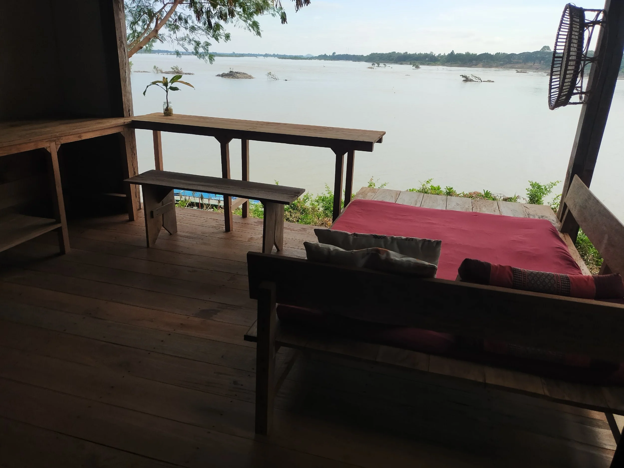 A rustic wooden porch overlooking a body of water with trees and small islands, featuring a wooden bench, a small table, and a daybed with a pink cushion and pillows.