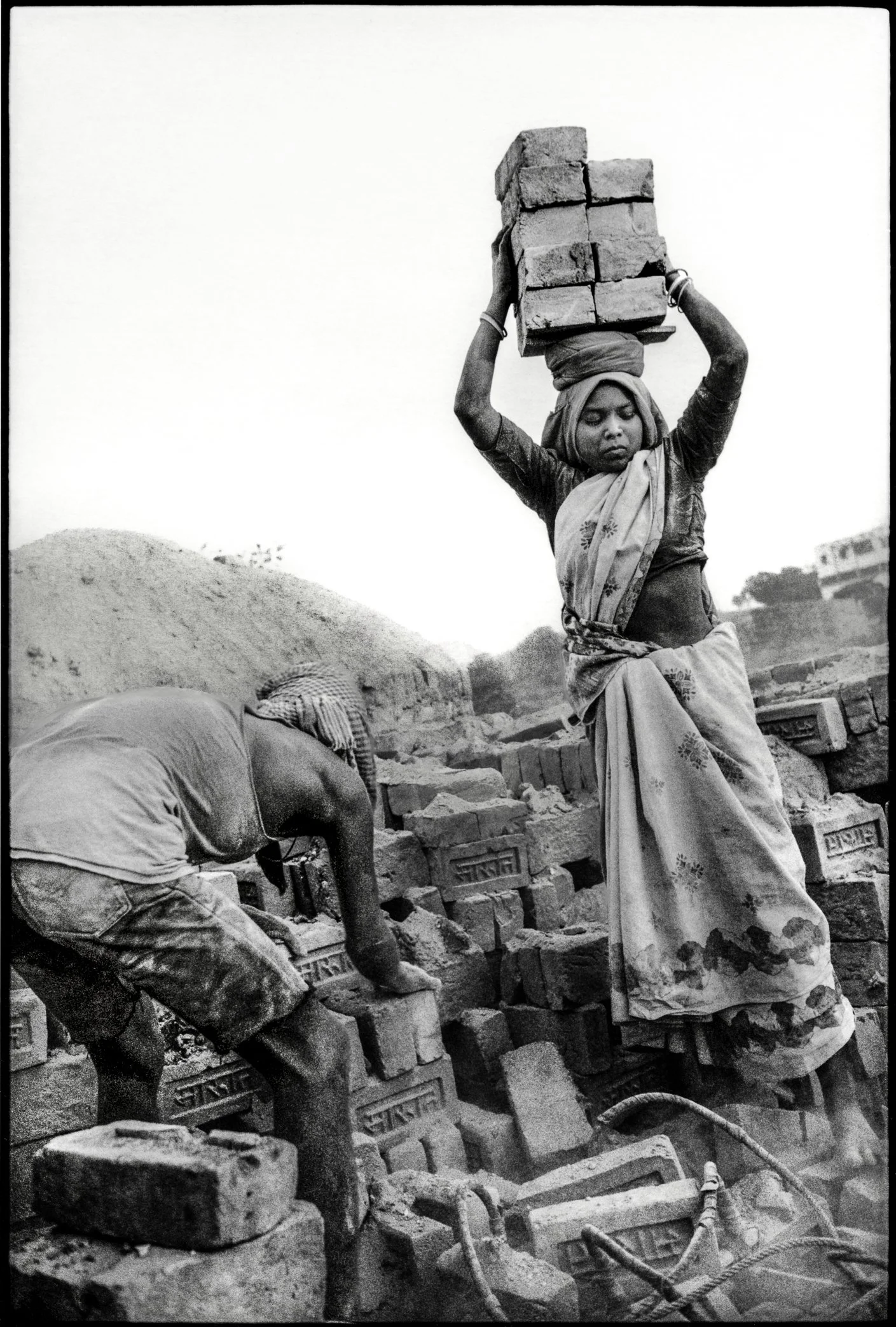 Patna, Bihar, India, April 2003 - Workers move freshly fired bricks from the kiln yard for transport and sale. Each load is carried by hand, through dust thick enough to become part of the work itself - entering the air, the skin, and the lungs of th