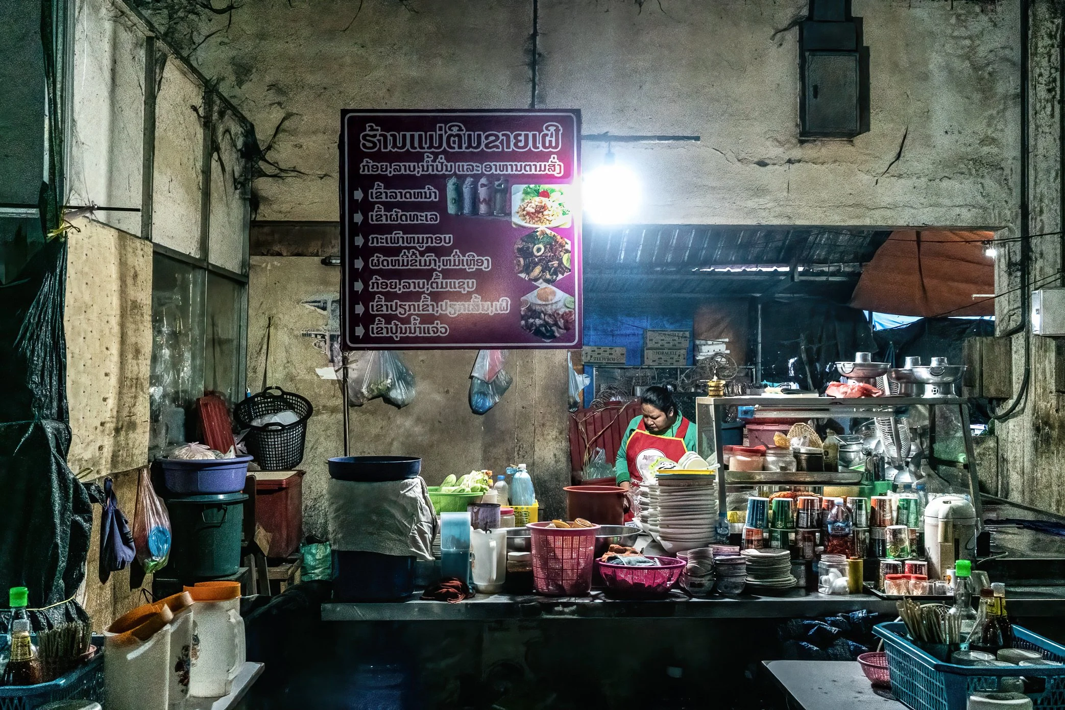 Pakse, Laos, 2022 - A food-stall owner prepares her kitchen before opening for the day. Nearly every seasoning and ingredient used in her shop comes from neighboring countries, tying even a small local meal to exchange rates, fuel costs, and the fall