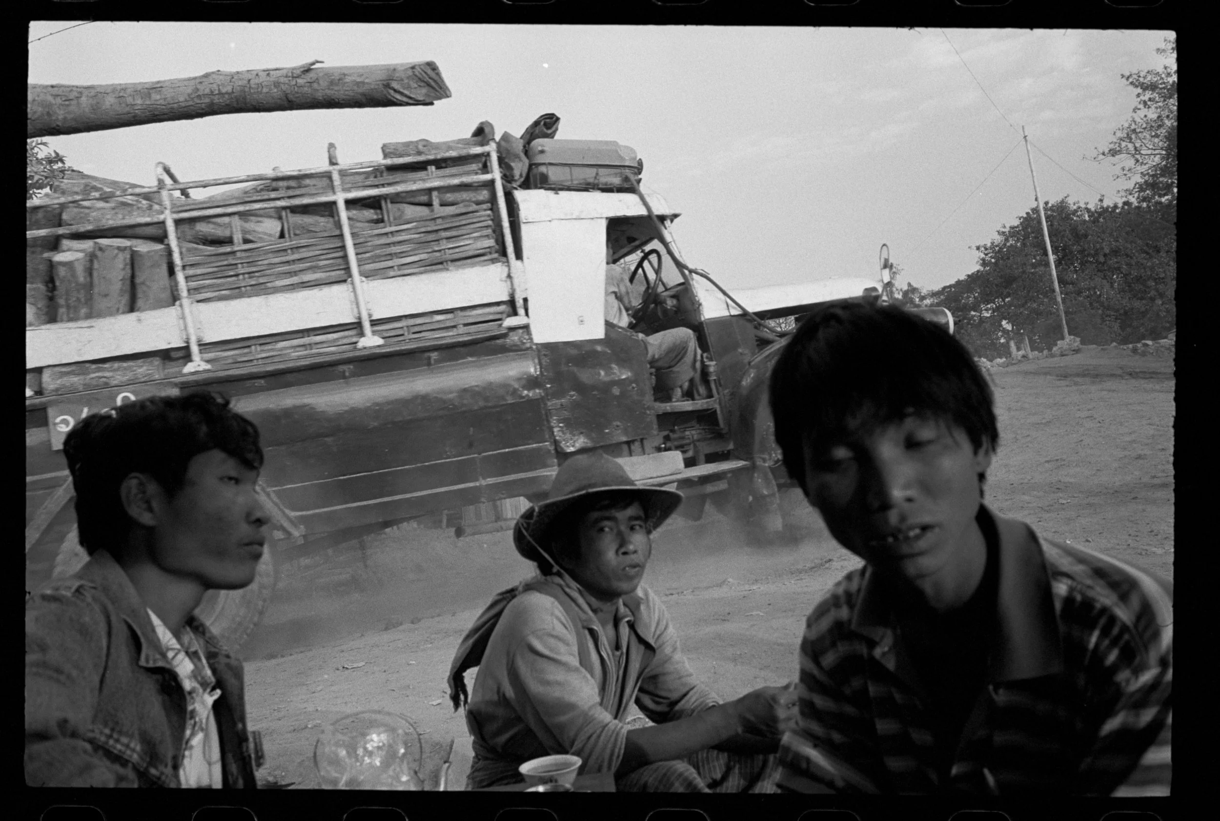 Three boys sitting outdoors with a truck in the background, which is loaded with logs and a large tree branch. One boy wears a hat, and the others have dark hair. The scene appears to be in a rural or semi-rural area with a dirt ground and trees in t