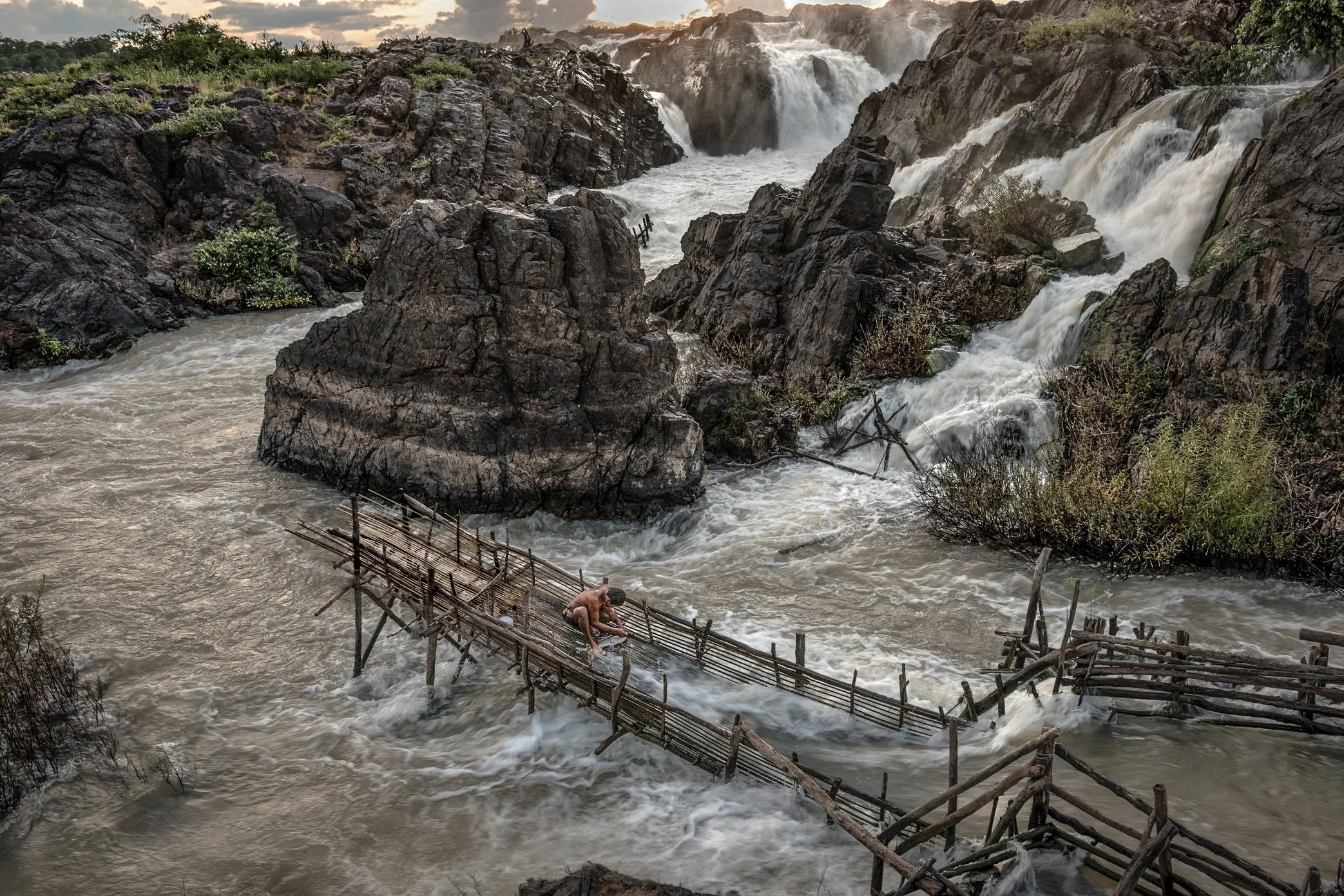 A fisherman checks his Li trap in a remote channel of Si Phan Don, where the Mekong breaks into waterfalls, rapids, and narrow passages between black rocks. Built by hand from bamboo, timber, and stone, the trap is placed where the current forces mig