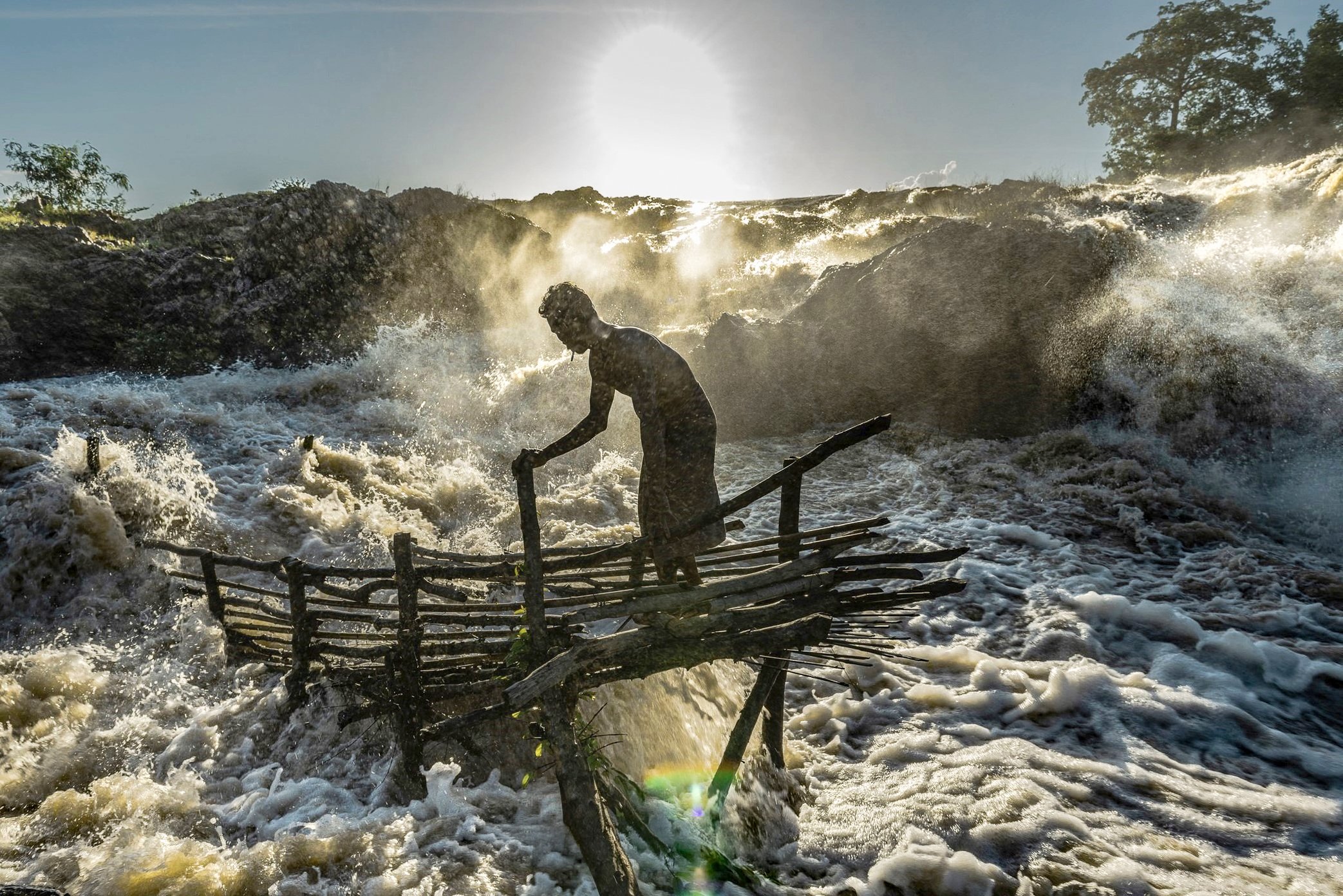 A fisherman checks his Li trap in the force of the midday current at Si Phan Don. During the migration season, the work is not only to collect fish, but to keep the trap alive against the river itself. Driftwood, branches, and debris carried by the M