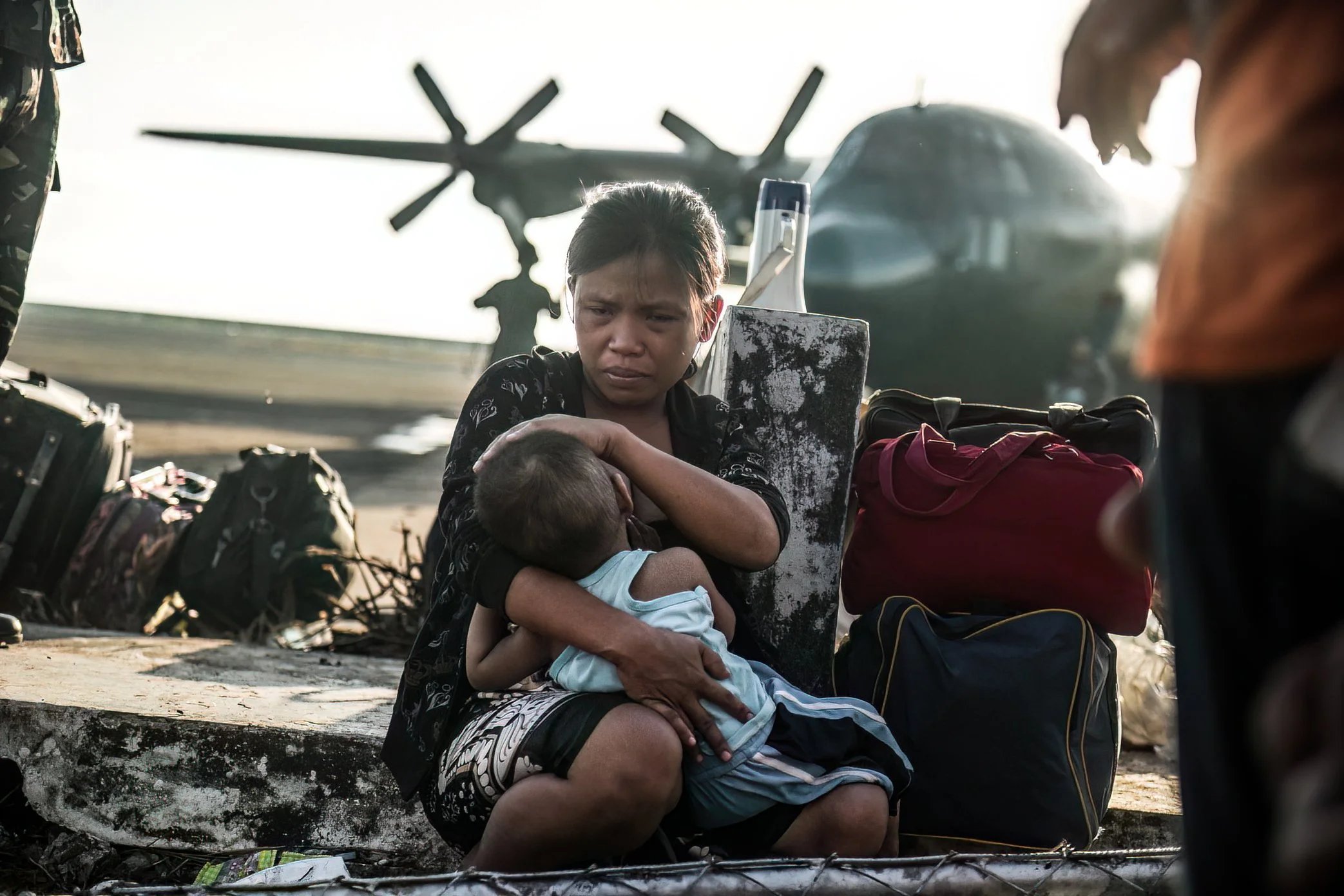 Tacloban, Philippines, 2013 - At Tacloban airport, a mother nurses her child while waiting for evacuation after Typhoon Haiyan. Around her, families sit with what they were able to carry from the ruins, hoping for a flight out of a city where surviva