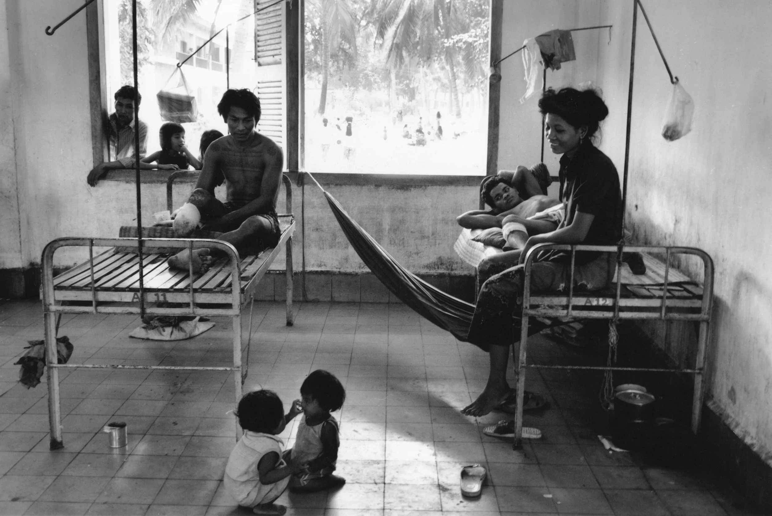 A black and white photo of a hospital room with two patients lying on beds, a woman sitting beside one bed, and children playing on the floor. There are people looking out the window.