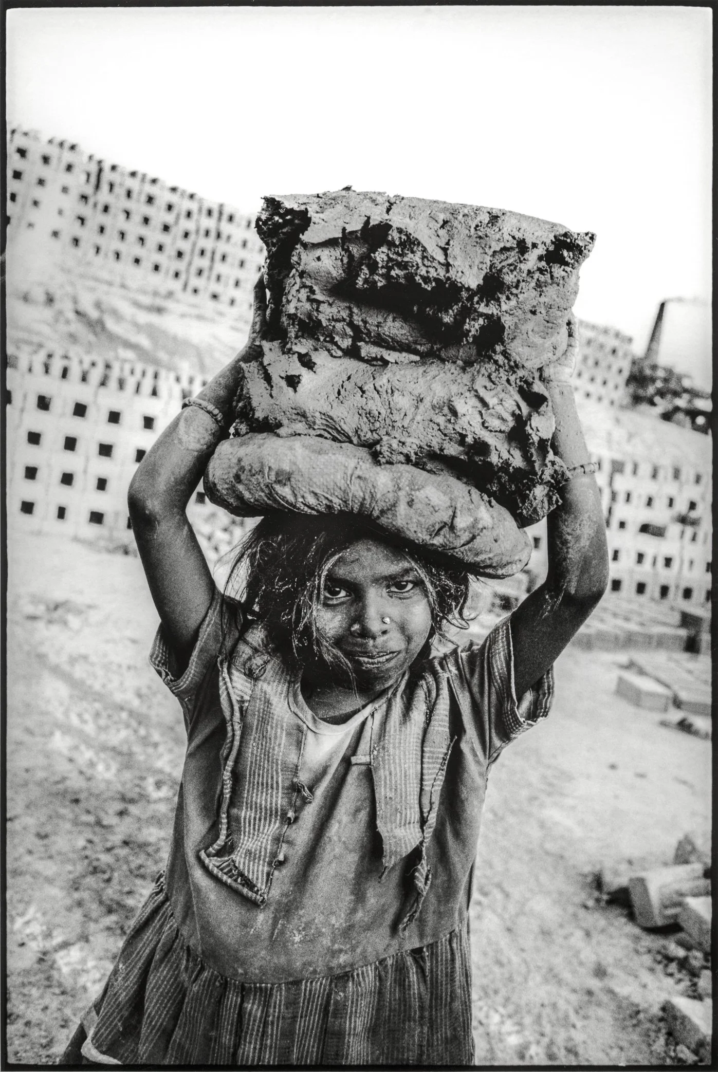 Patna, Bihar, India, April 2003 - A young girl carries freshly mixed clay on her head at a brick kiln in Bihar. Around her, walls of drying bricks rise from the earth, while childhood is folded into the same cycle of labor as the adults beside her.