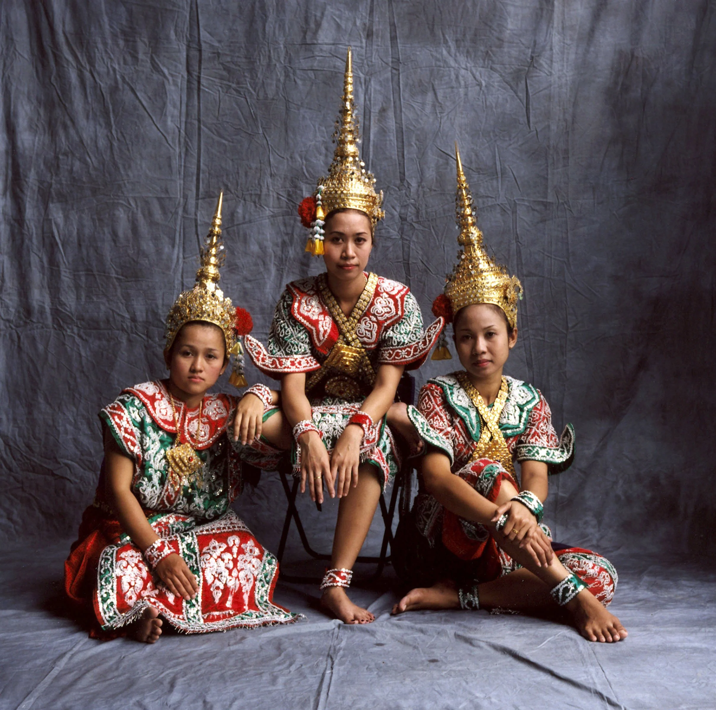 Three women dressed in traditional Thai costumes with intricate gold headpieces, sitting and kneeling on a gray backdrop.
