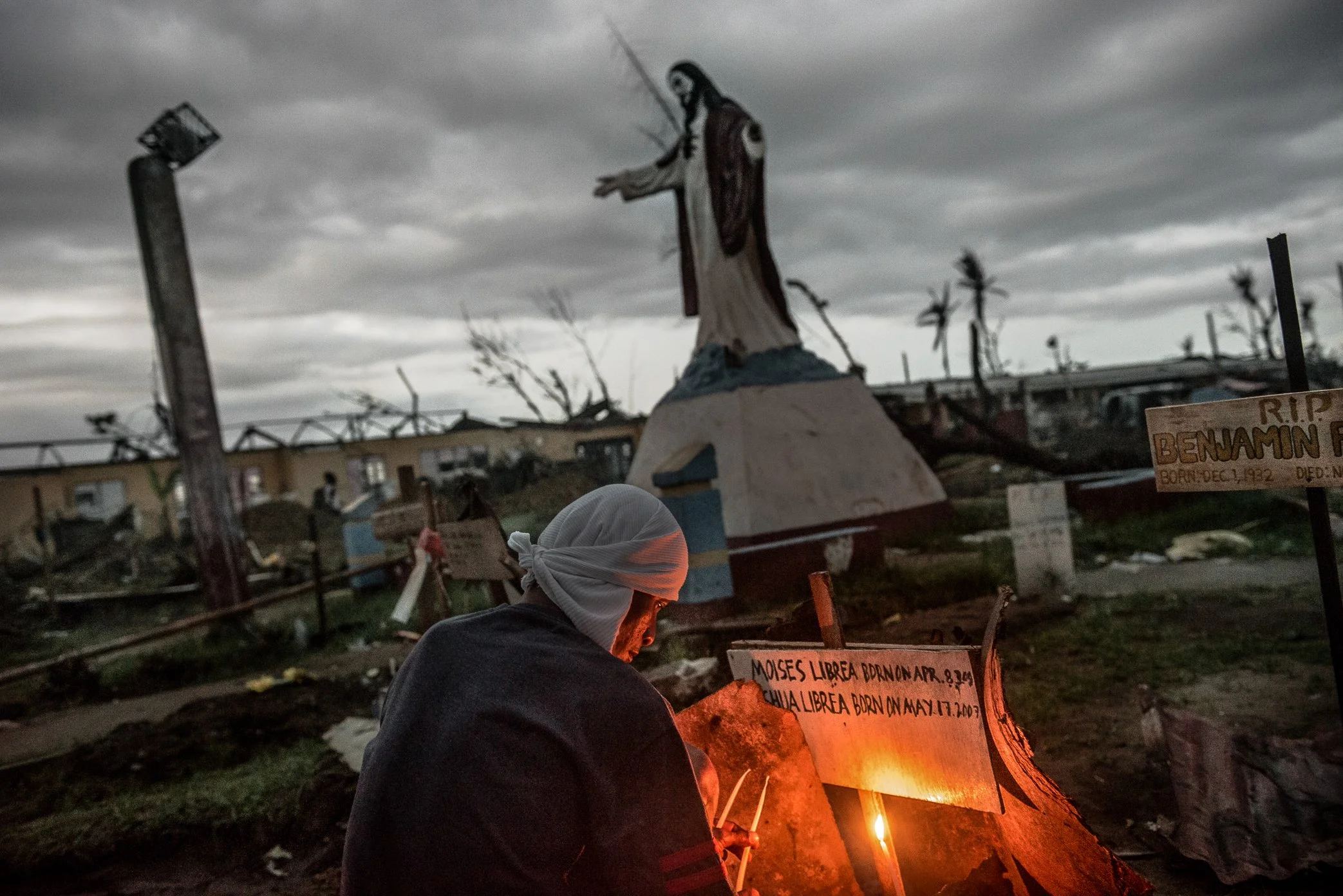 Palo, Leyte, Philippines, 2013 - At dusk, candles burn in a makeshift graveyard for victims of Typhoon Haiyan. Some graves mark entire families; in some homes, no one survived to mourn them. Behind the candles, broken trees and damaged buildings rema
