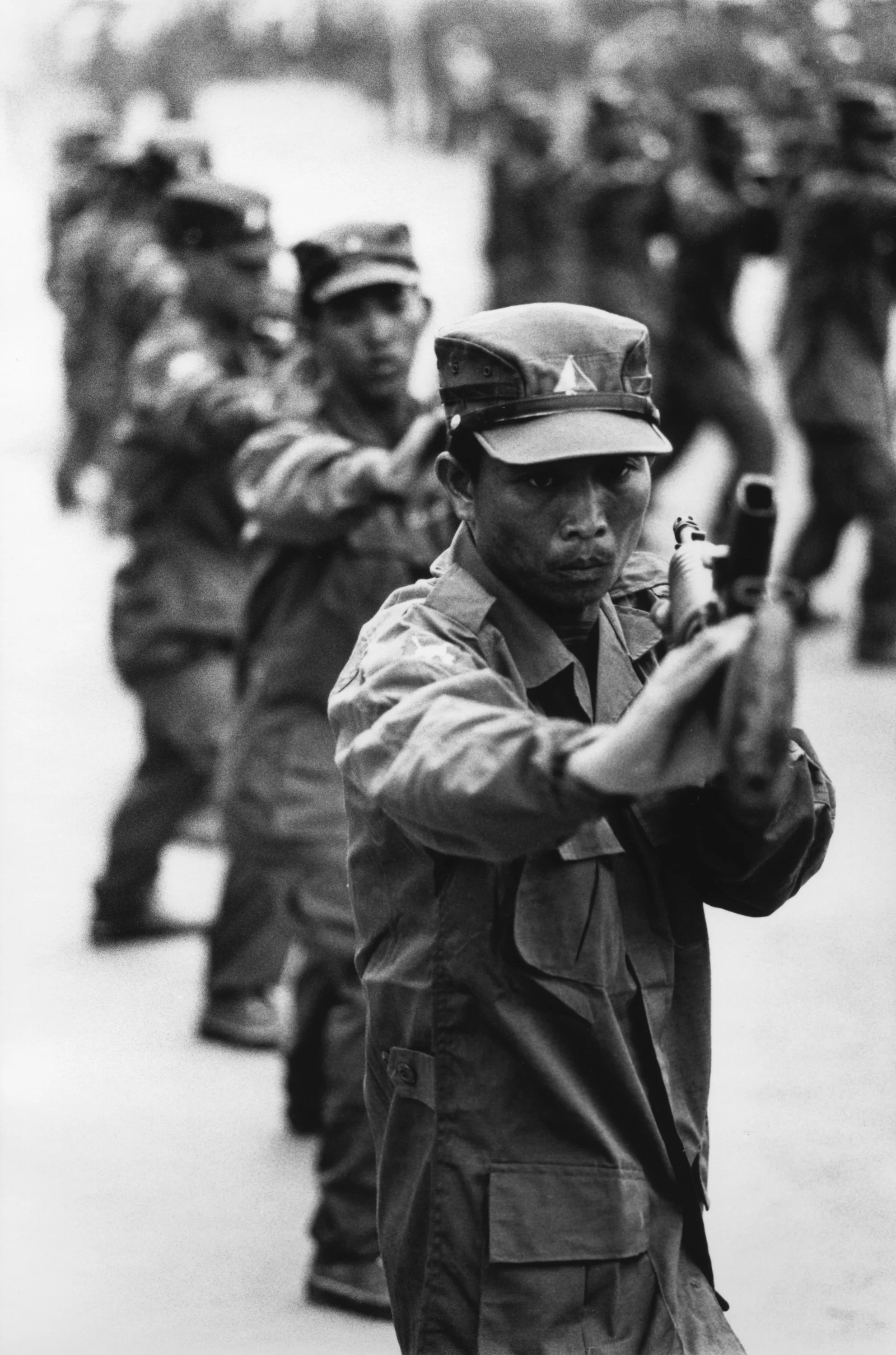 A black and white photo of soldiers in uniform, with the soldier in the foreground aiming a gun directly at the camera, amidst a line of soldiers in the background.
