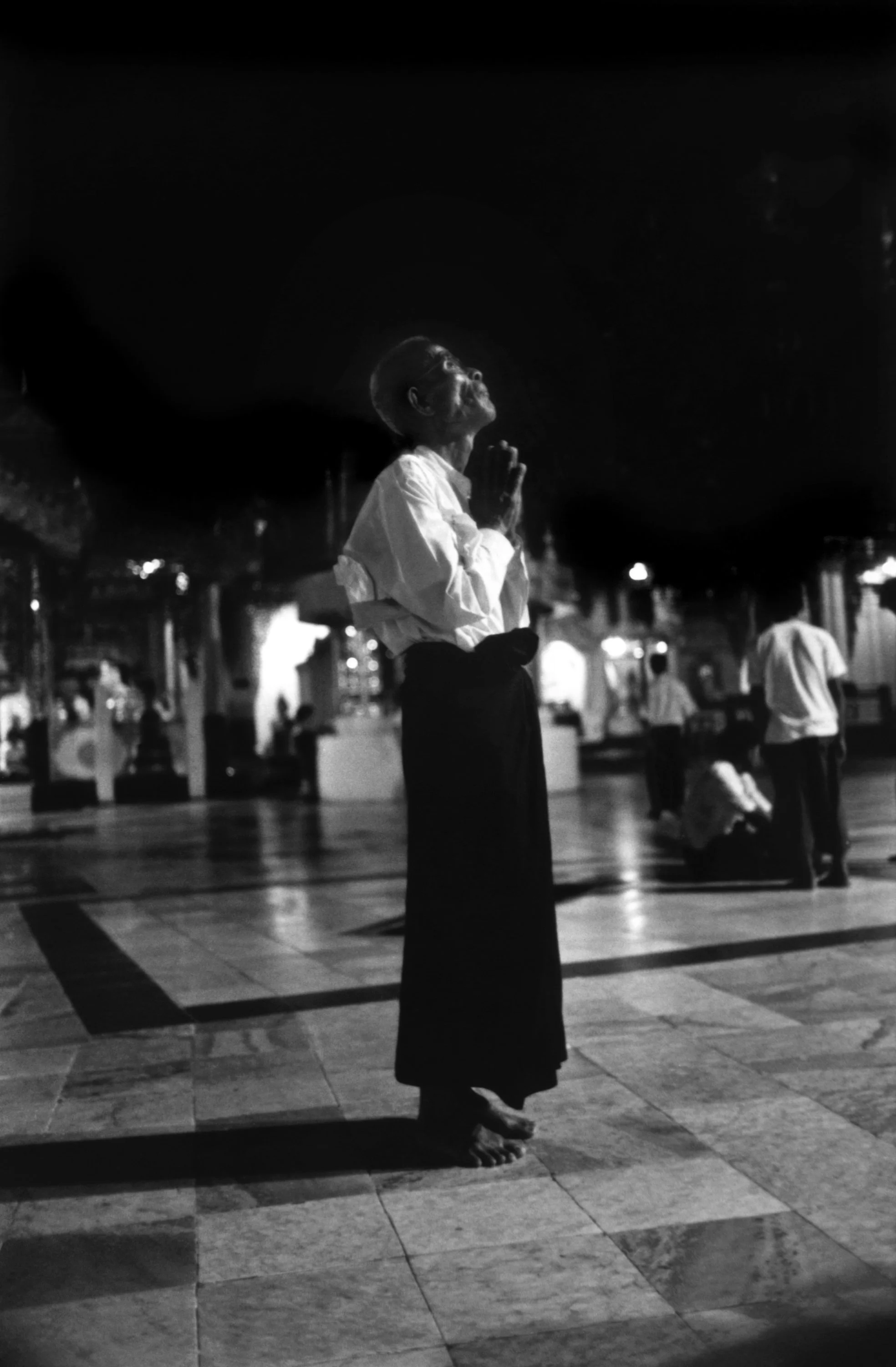A person standing barefoot on a tiled floor, praying with hands clasped and head tilted back in a dimly lit indoor space, with other people in the background.