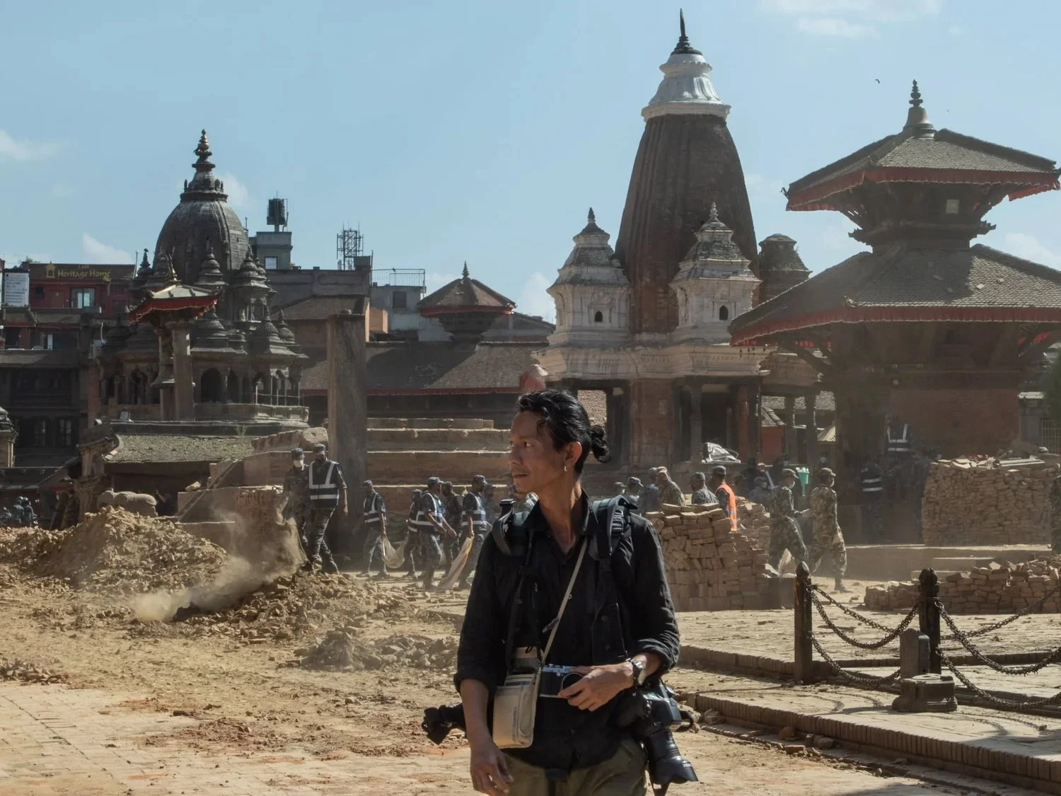 A woman holds a camera in front of ancient temples and ruins, with construction activity and workers in the background, in a historical or archaeological site.