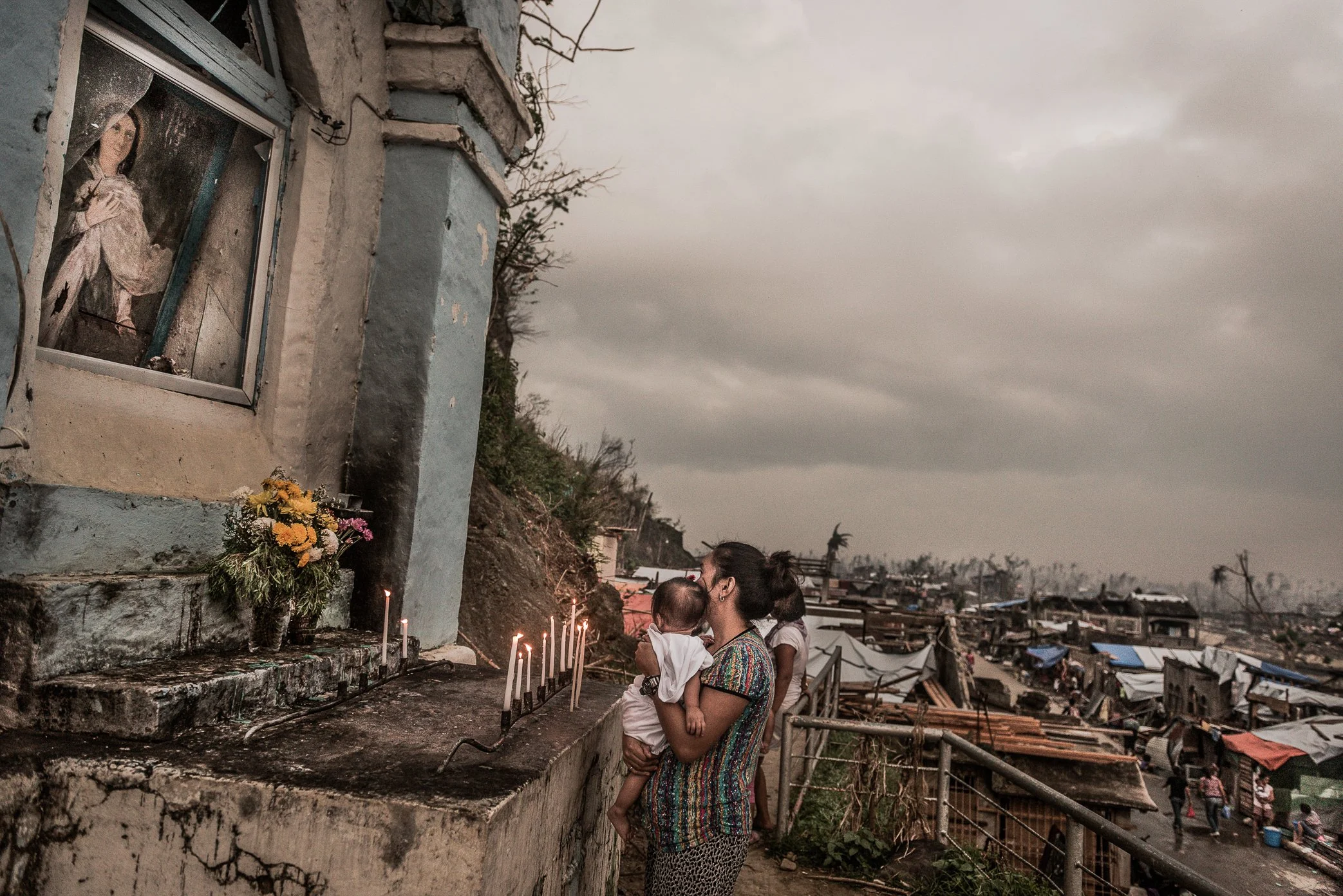 Palo, Leyte, Philippines, 2013 - At a hillside shrine overlooking a town scarred by Typhoon Haiyan, a young mother holds her child and lights candles in prayer. Below them, families rebuild among damaged homes and broken trees, while faith becomes on