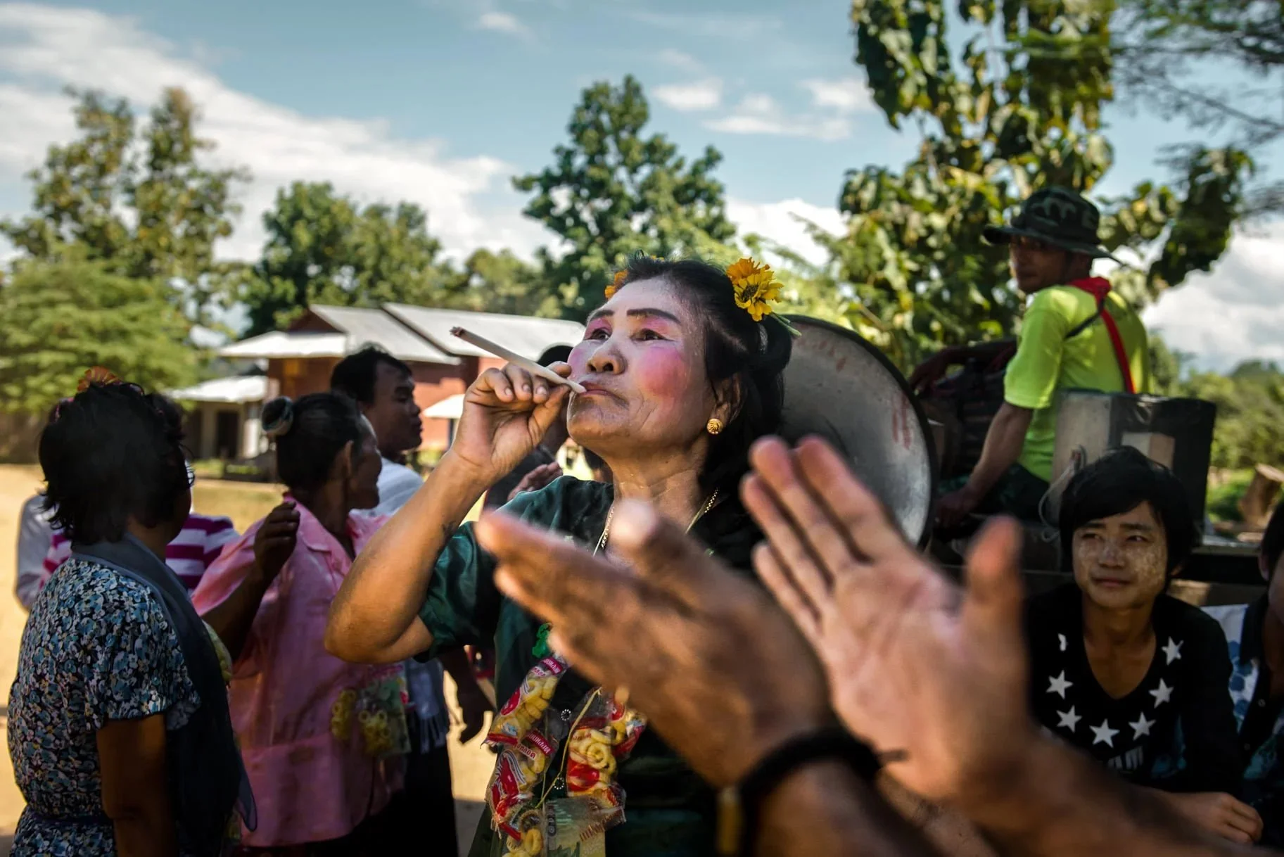 A woman dressed in traditional attire, with floral decorations in her hair, is smoking a cigarette in a rural outdoor setting with a group of people surrounding her and trees in the background.