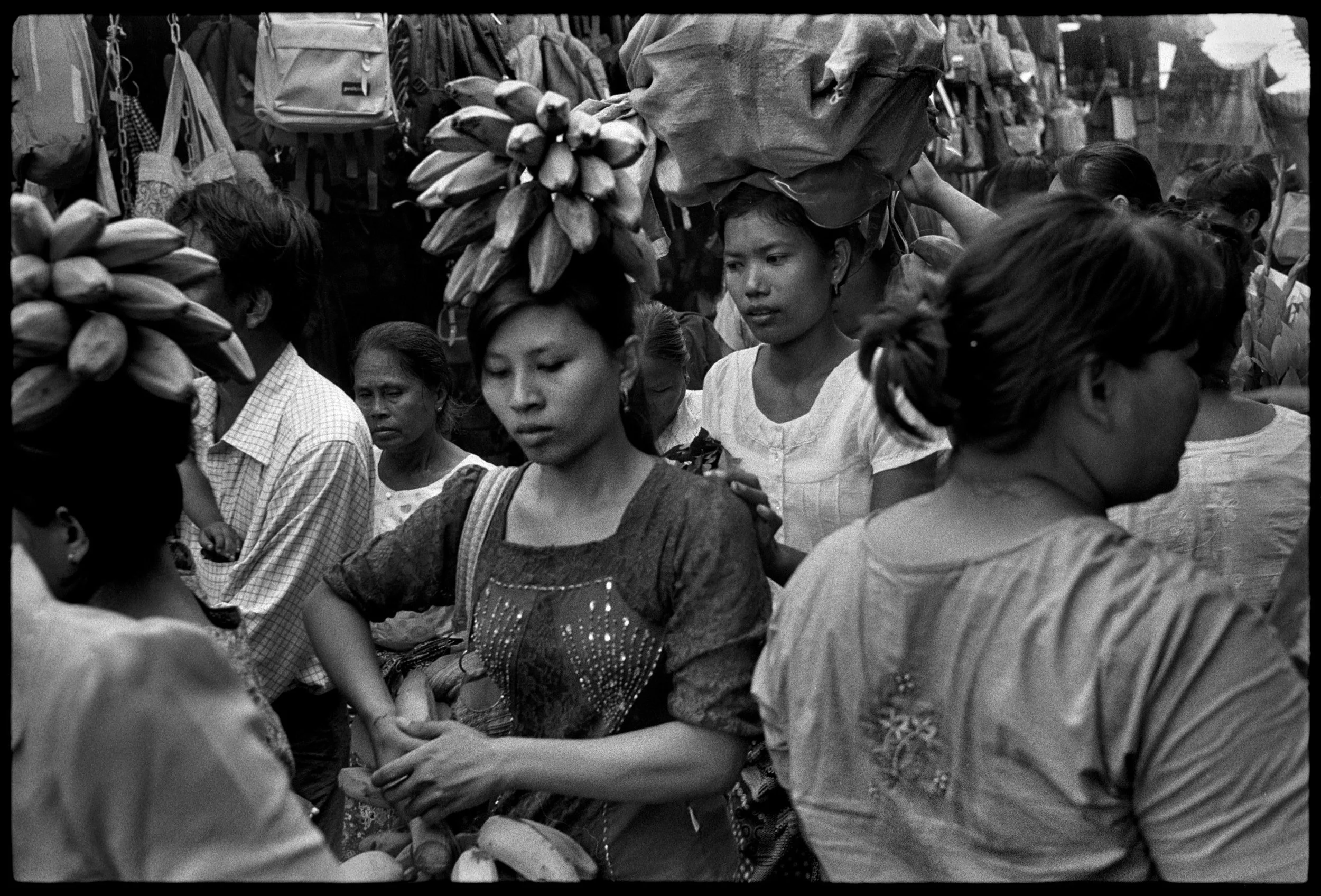 Black and white photograph of a crowded marketplace with women shopping. Some women carry large bundles of bananas on their heads, while others hold items in their hands. The background shows hanging bags and more shoppers.