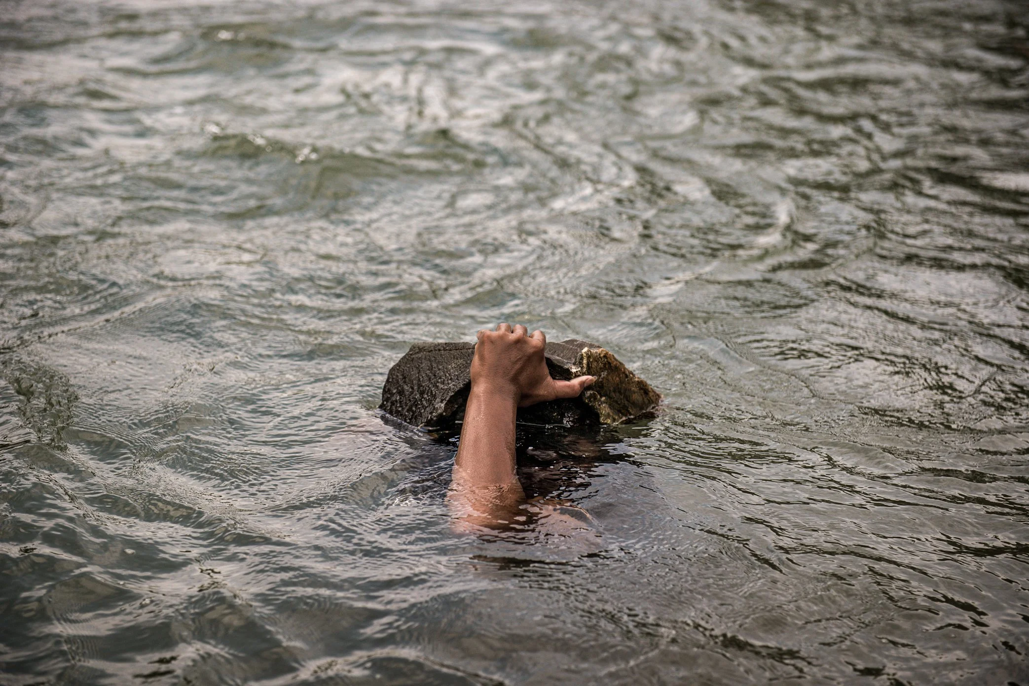 A fisherman dives for stones to build the foundation of a Li trap in Si Phan Don. Throughout the day, men search underwater, lifting rocks by hand and placing them at the base of the wooden frame. The weight is essential: if the foundation is too lig